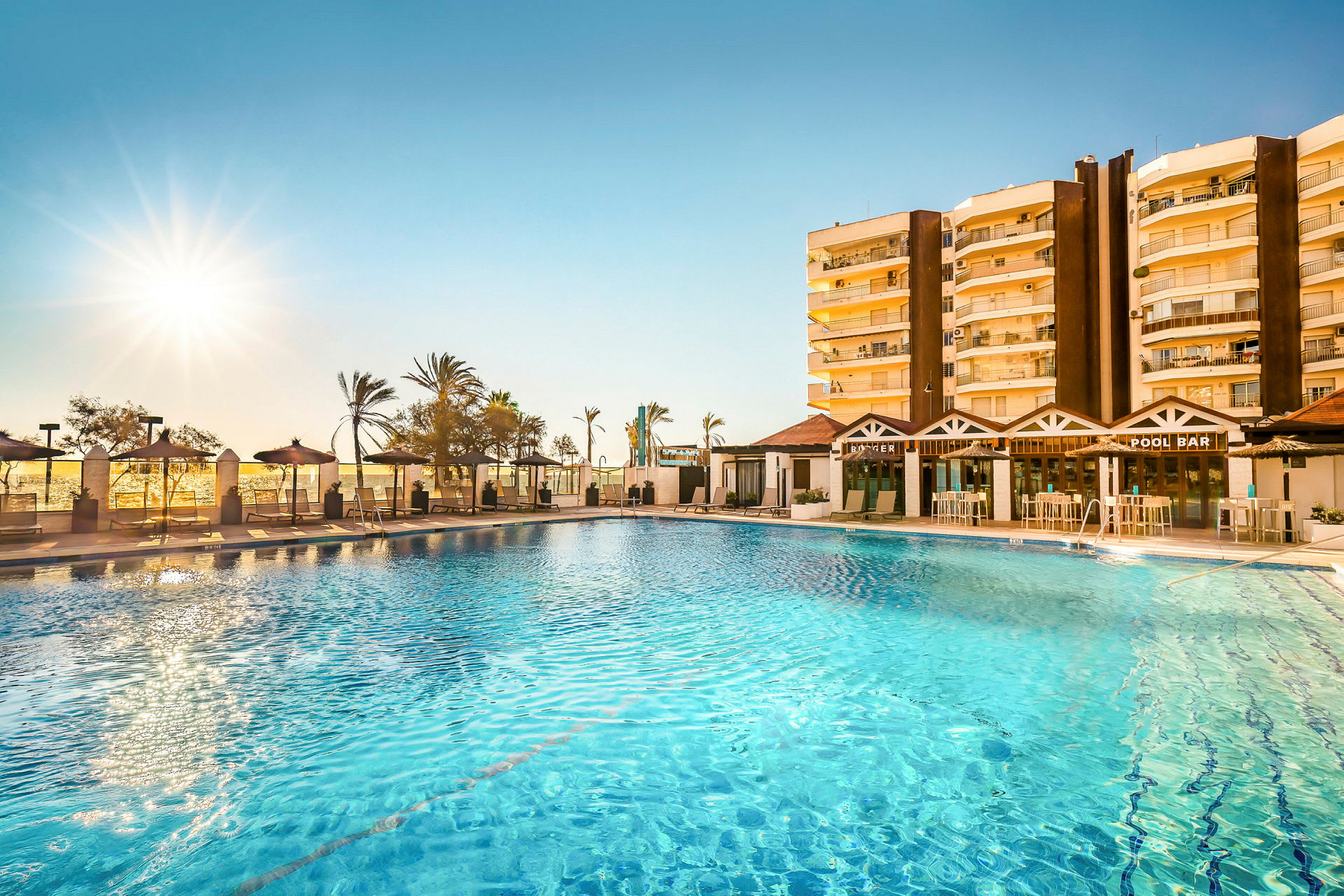 Bild av en hotellpool med barområde och solstolar, palmträd och ett hotell i bakgrunden. (AI-genererad text)