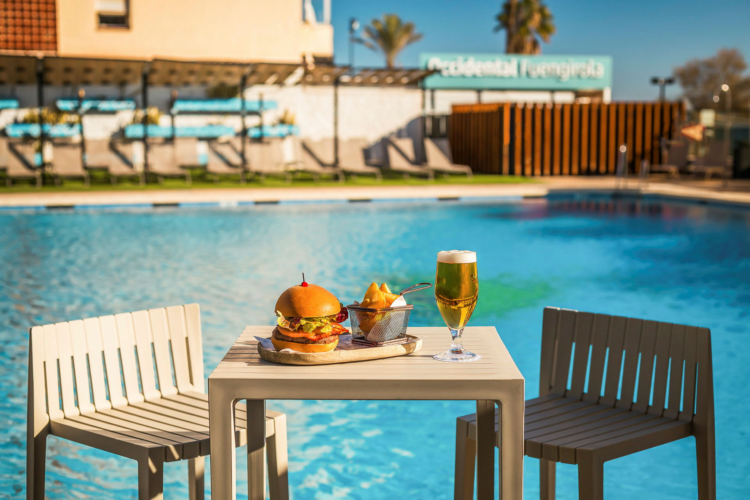 Ett bord med en hamburger tallrik och en ölglas bredvid en pool. (AI-genererad text)