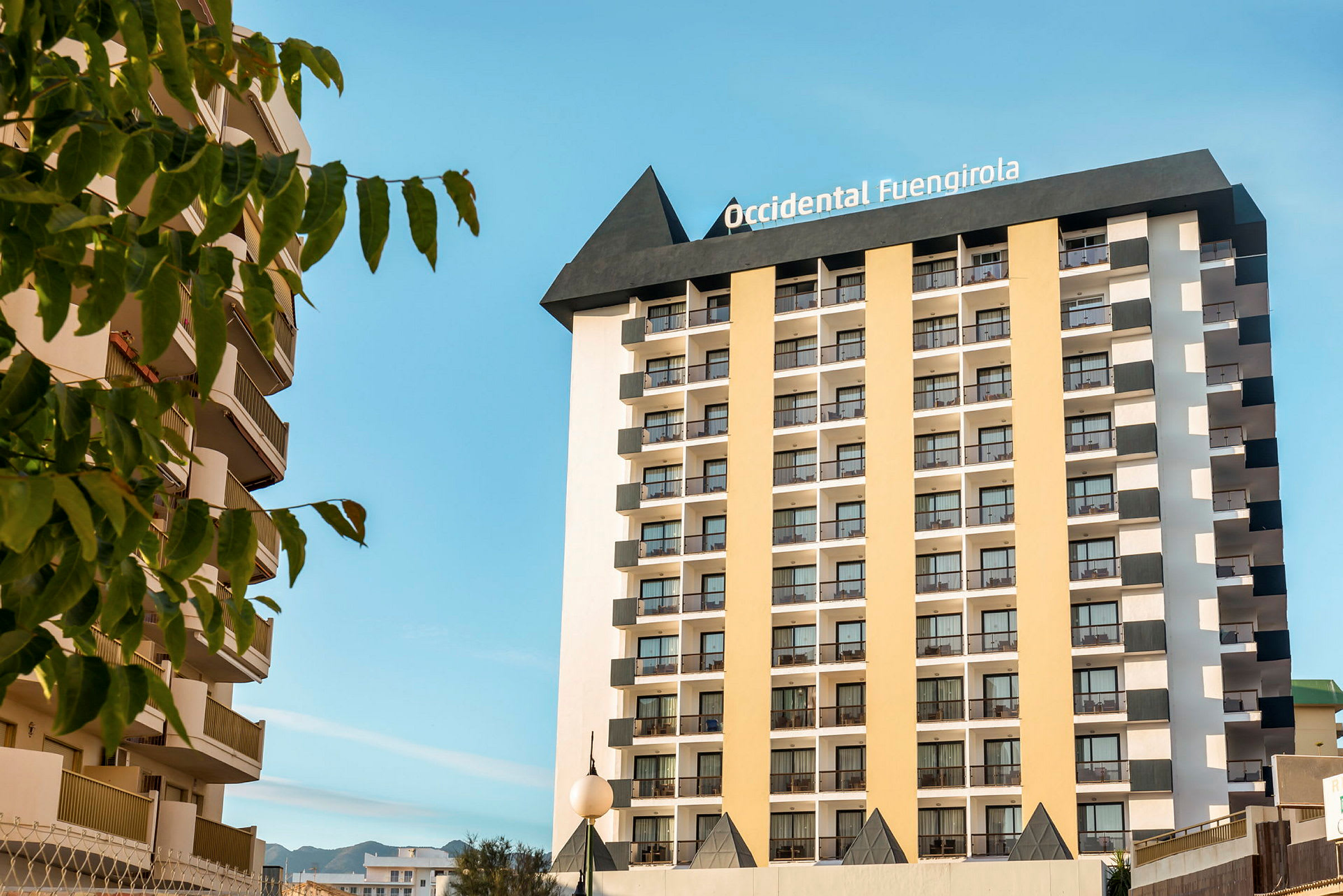 Bild av ett hotell med många våningar och balkonger, omgivna av träd och under en klarblå himmel. (AI-genererad text)