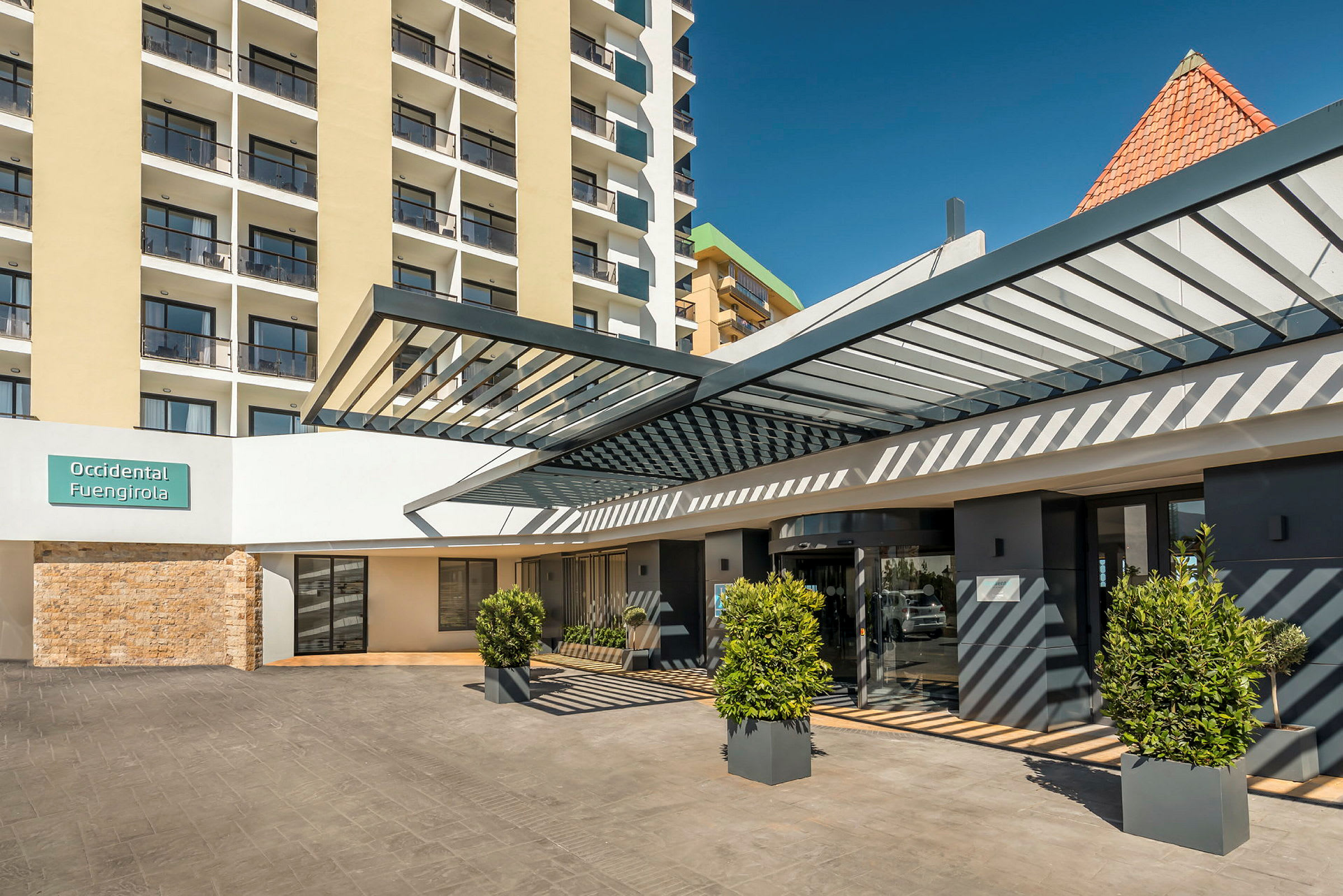 Entrén till en modern byggnad med skylt som lyder 'Occidental Fuengirola', omgiven av planterade buskar och en pergolaliknande konstruktion. (AI-genererad text)