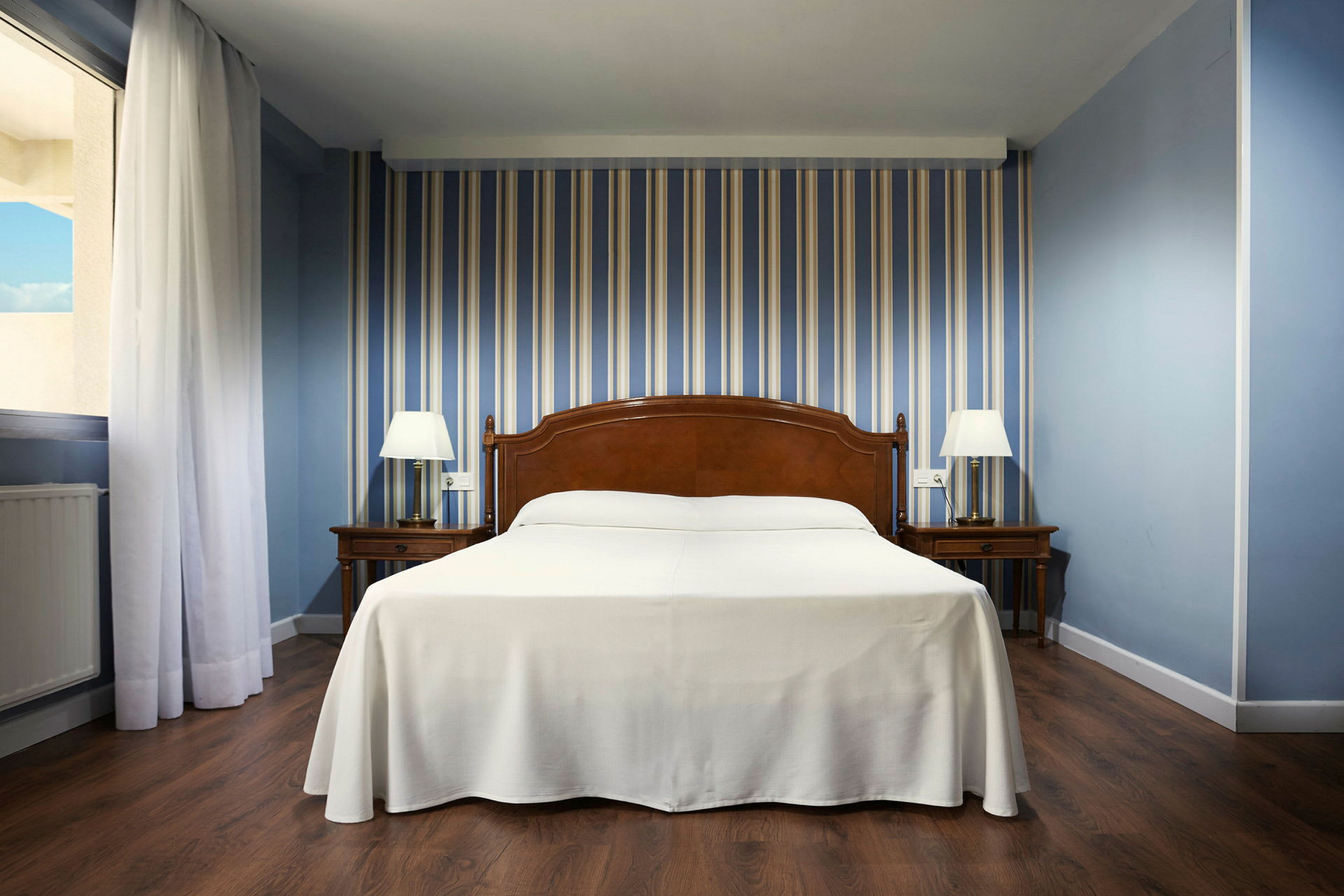 Hotellrum med säng i mitten, två sängbord med lampor och blårandig tapet. (AI-genererad text)