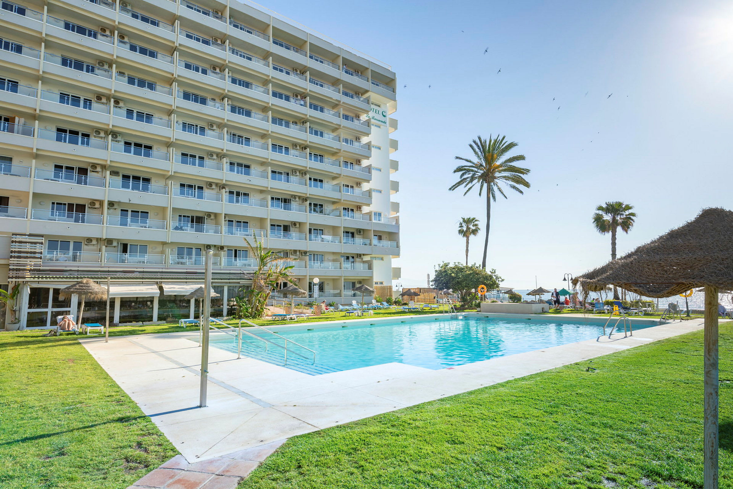 Hotellets poolområde med gröna gräsmattor, solparasoller och en byggnad i bakgrunden. (AI-genererad text)