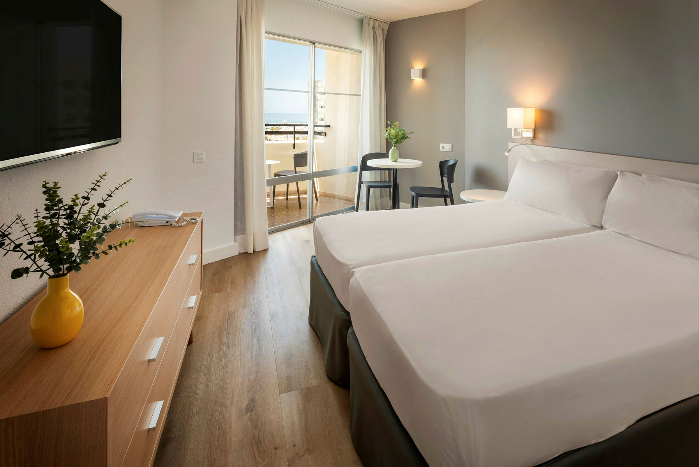 Ett hotellrum med två sängar, ett träbord med en gul vas och en balkong i bakgrunden. (AI-genererad text)