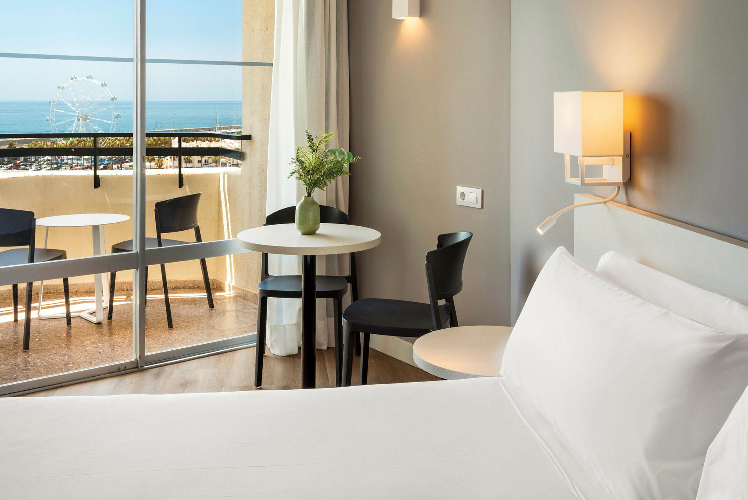 Ett hotellrum med en säng, bord och stolar nära en balkong med utsikt över havet. (AI-genererad text)