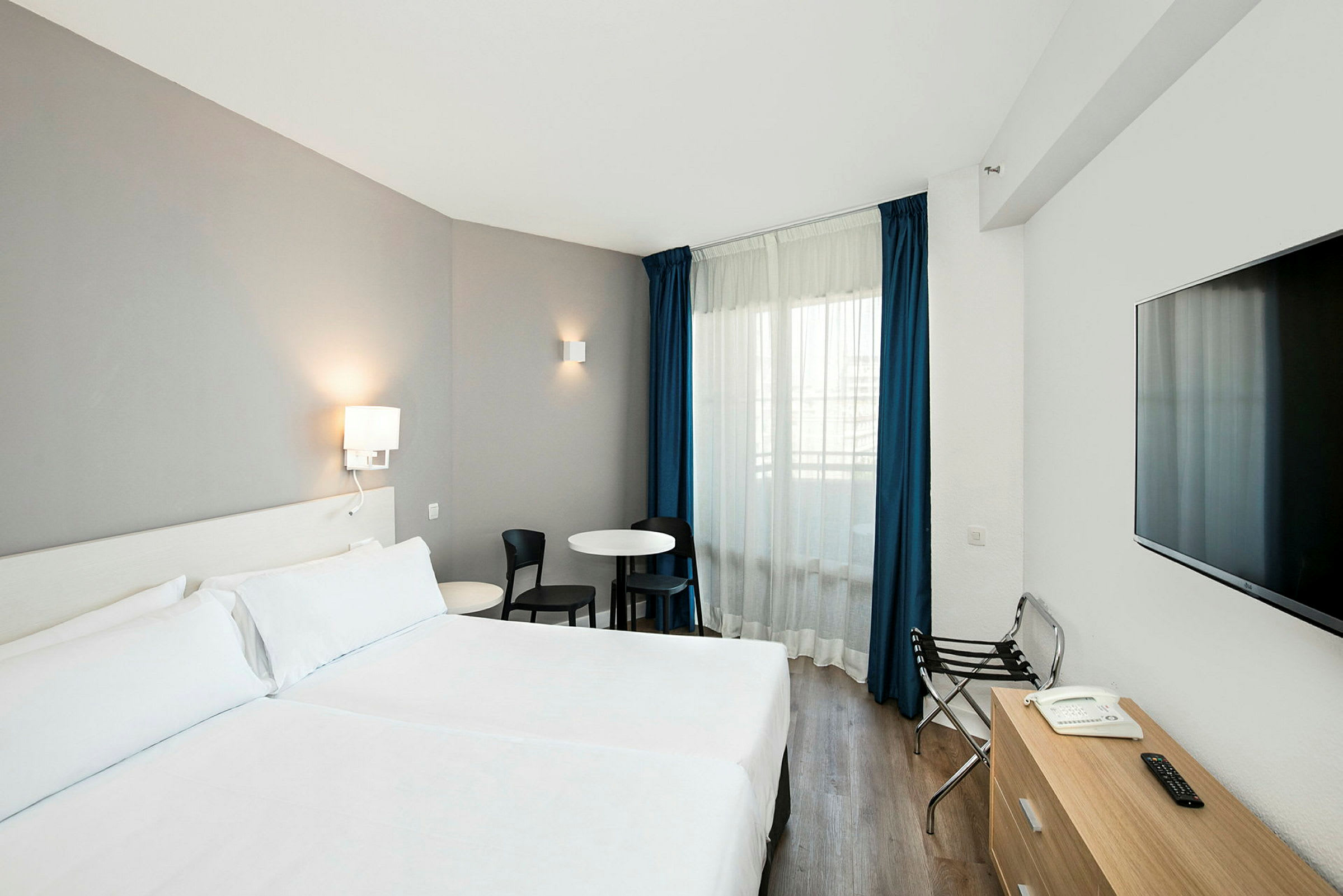 Ett hotellrum med en dubbelsäng, ett skrivbord, en stol och ett fönster med blå gardiner. (AI-genererad text)