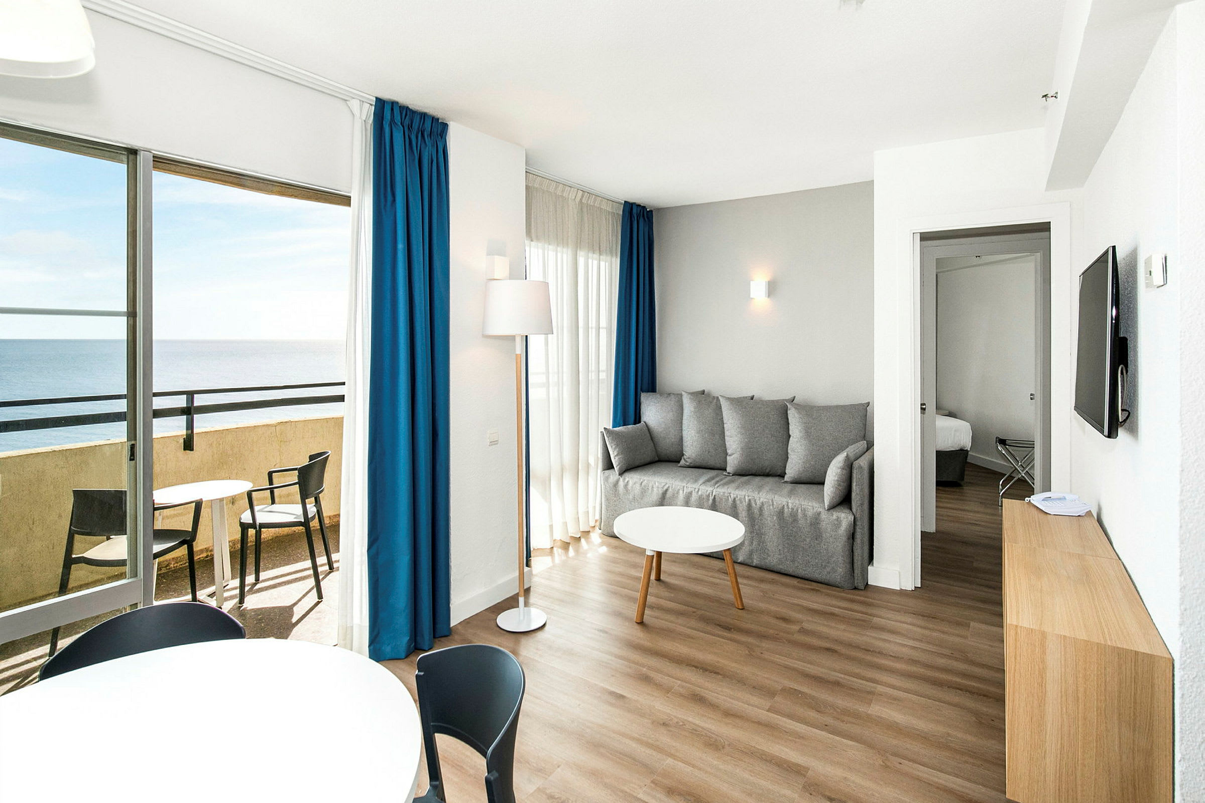 Ett vardagsrum med en grå soffa, ett runt soffbord och ett balkongområde med havsutsikt. (AI-genererad text)