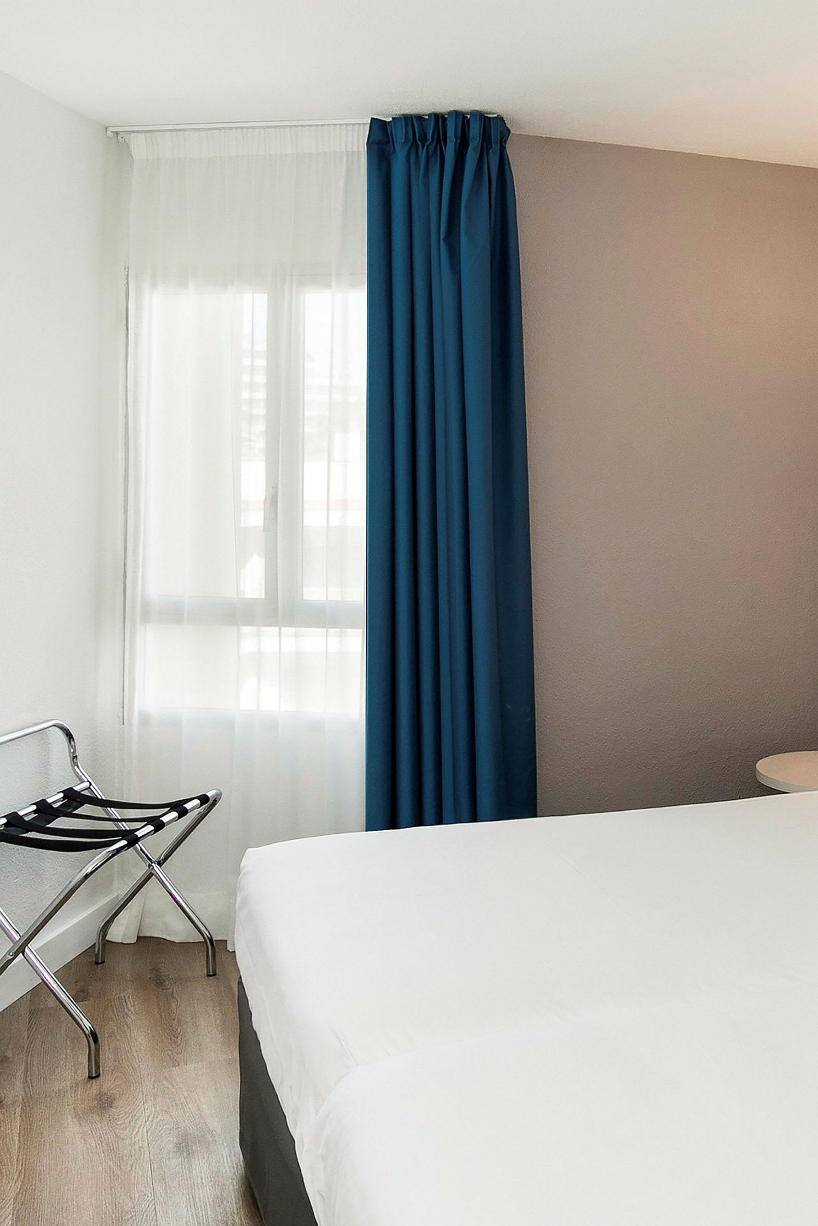 Ett hotellrum med ett vitt sängöverdrag, en blå gardin och en bagagehållare vid sidan. (AI-genererad text)