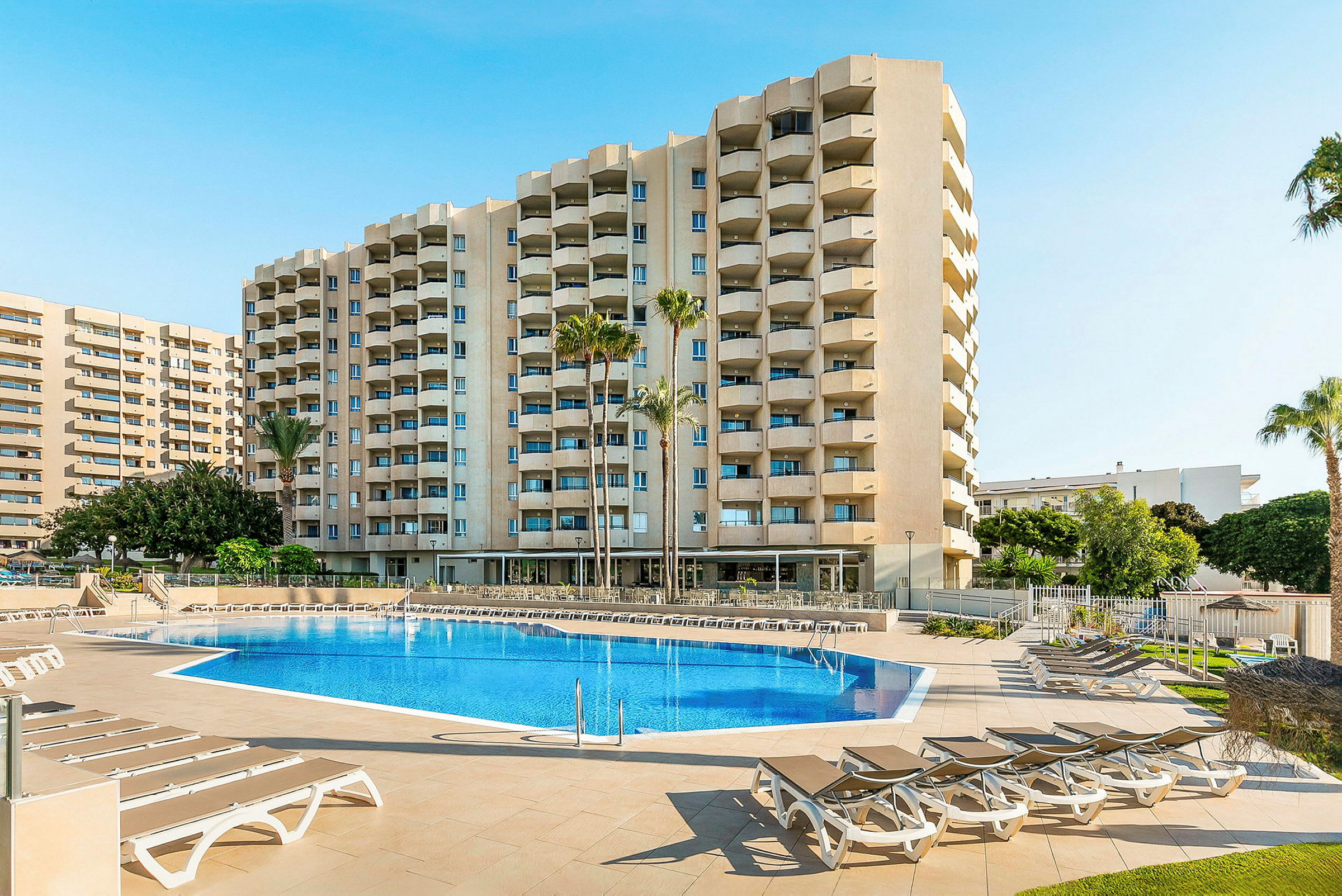 Hotellbyggnad med pool och solstolar i förgrunden. (AI-genererad text)
