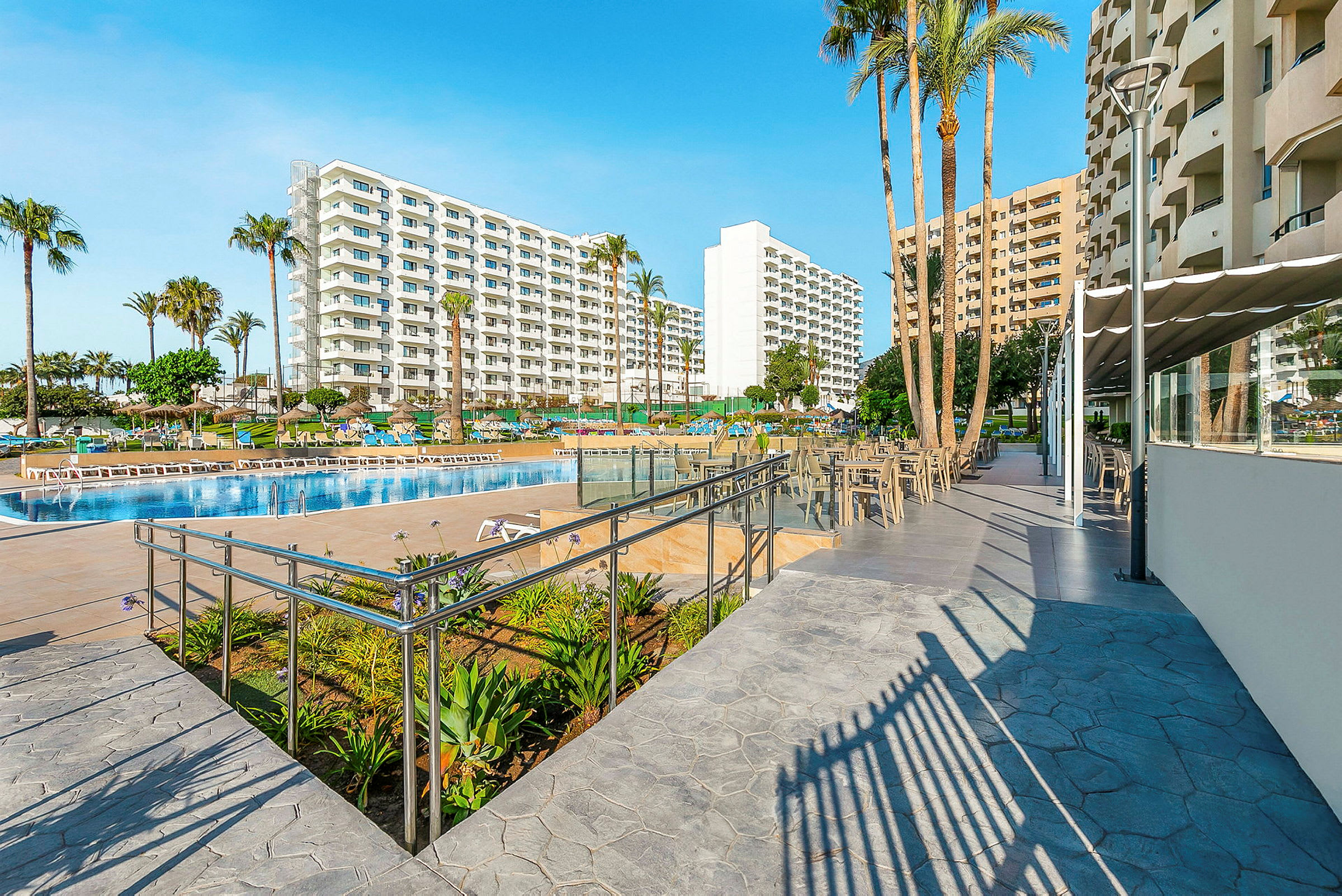 Utsikt över en hotellpool och omgivande byggnader med solstolar och palmer. (AI-genererad text)