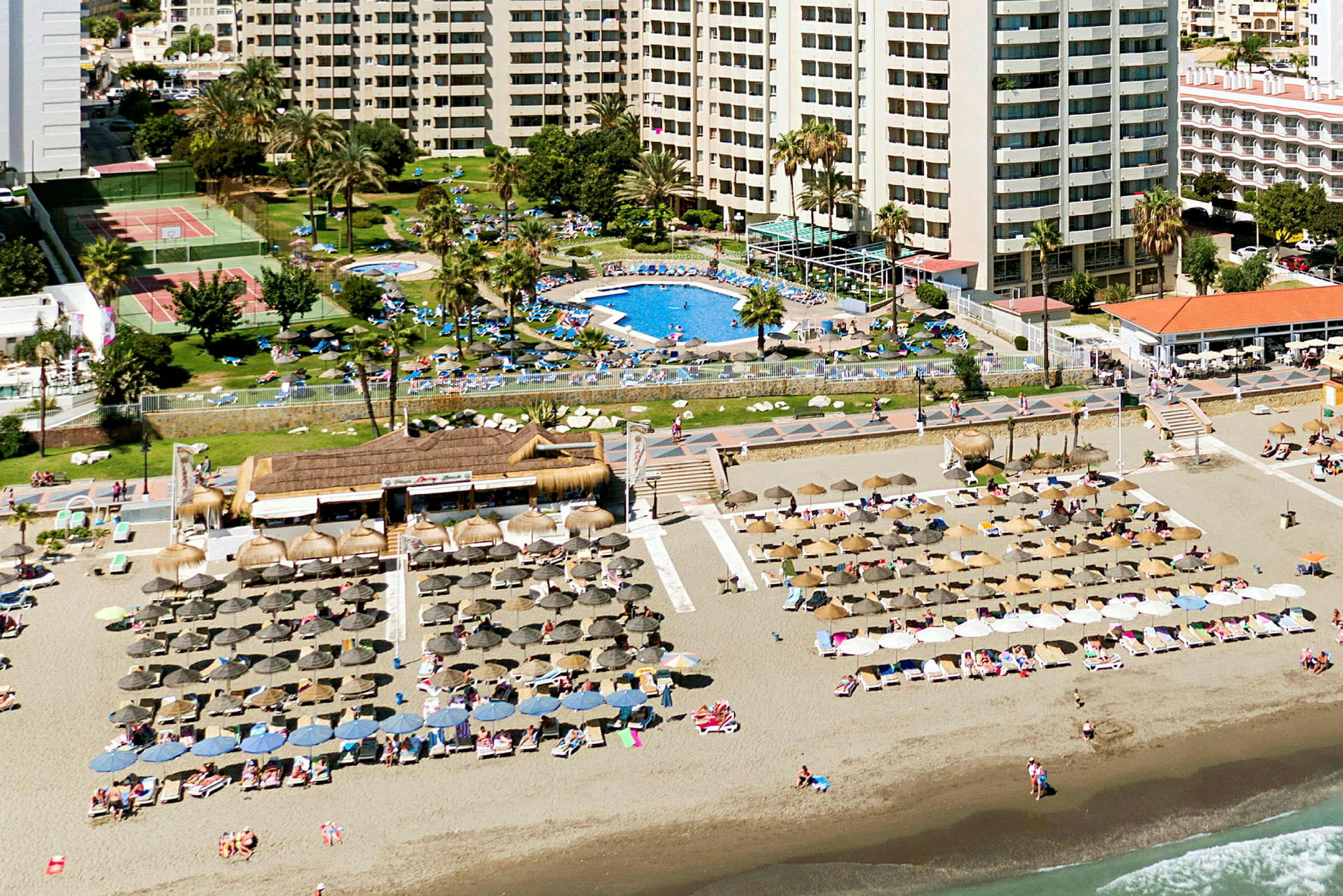 Bild av stranden med parasoller och solstolar framför hotellbyggnader och en poolområde. (AI-genererad text)