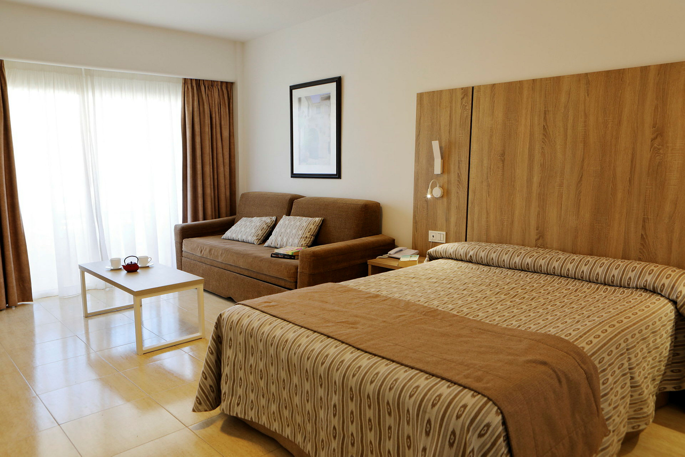 Hotellrum med en brun soffa, vita gardiner, ett bord och en säng. (AI-genererad text)
