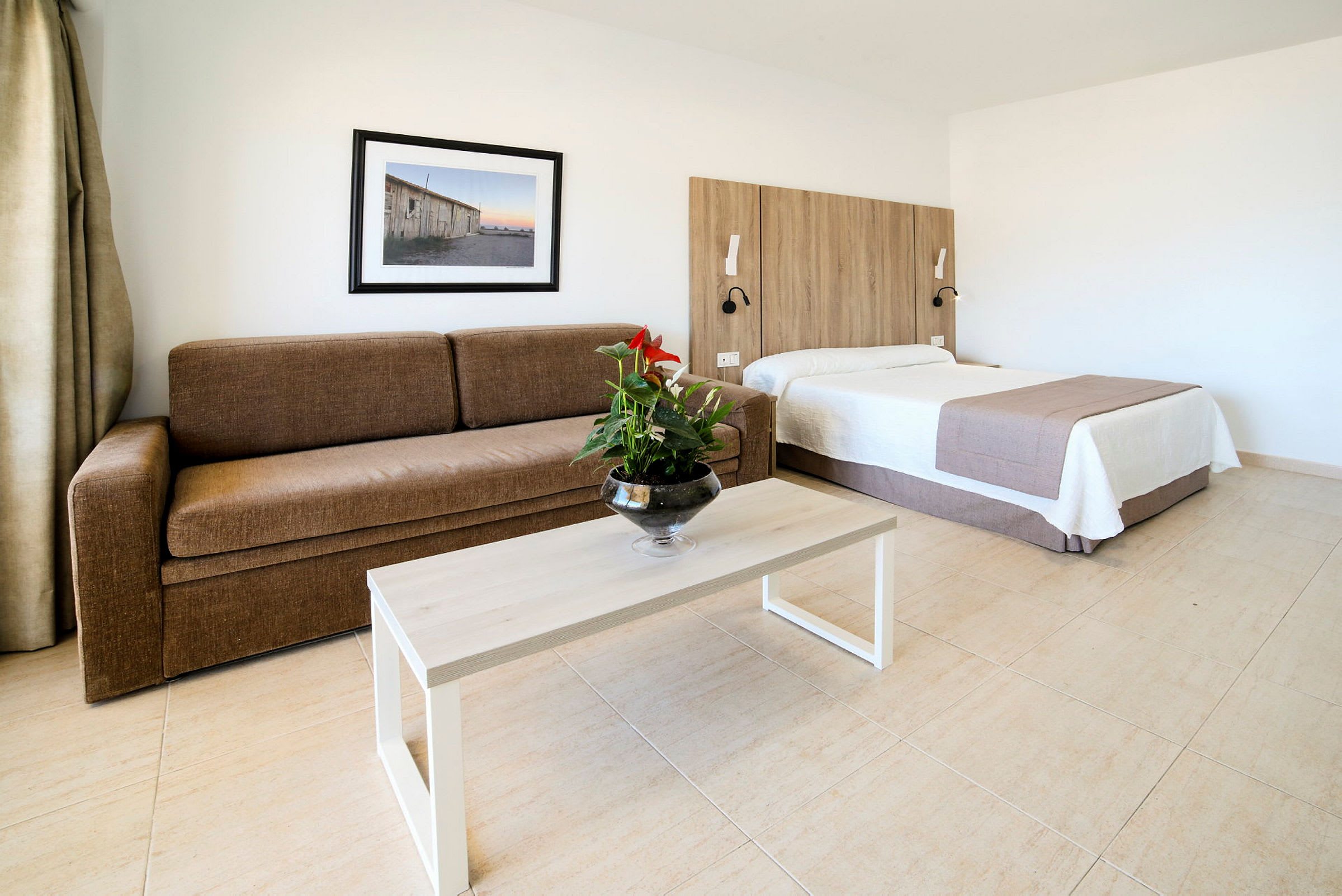 Ett hotellrum med en brun soffa, ett bord med en blomvas, och en säng med träpanel bakom. (AI-genererad text)