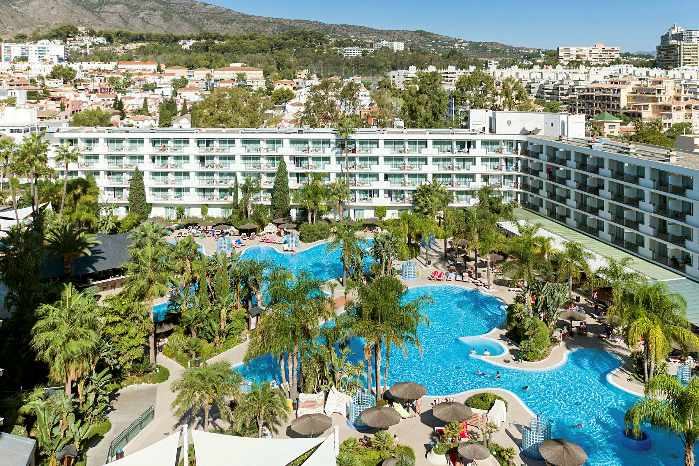 Ett hotellområde med pool, solstolar och palmer, omgiven av flerbostadshus och berg i bakgrunden. (AI-genererad text)