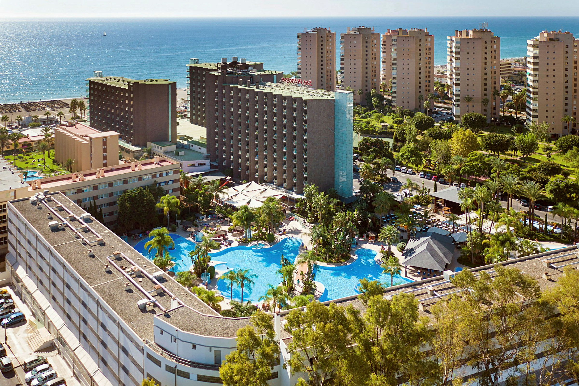 Flygfoto av en semesterort med hotellbyggnader, poolområde, trädgårdar och närliggande strand samt hav. (AI-genererad text)