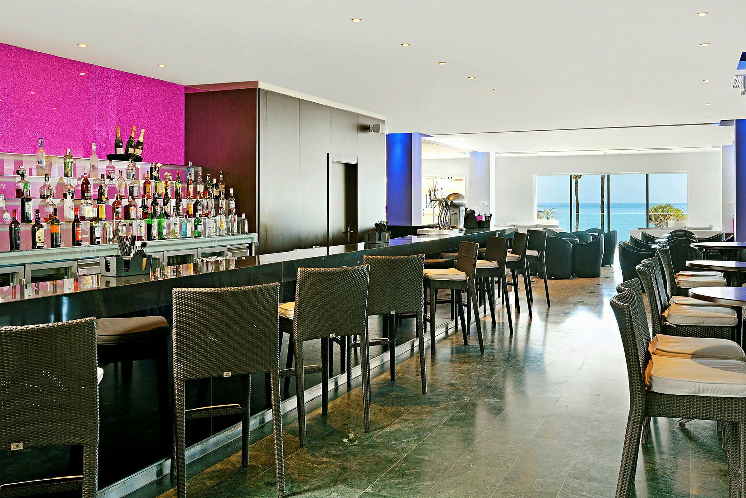 Bild av en modern bar med höga stolar, bord och olika färgglada lådor av sprit samt utsikt mot havet. (AI-genererad text)