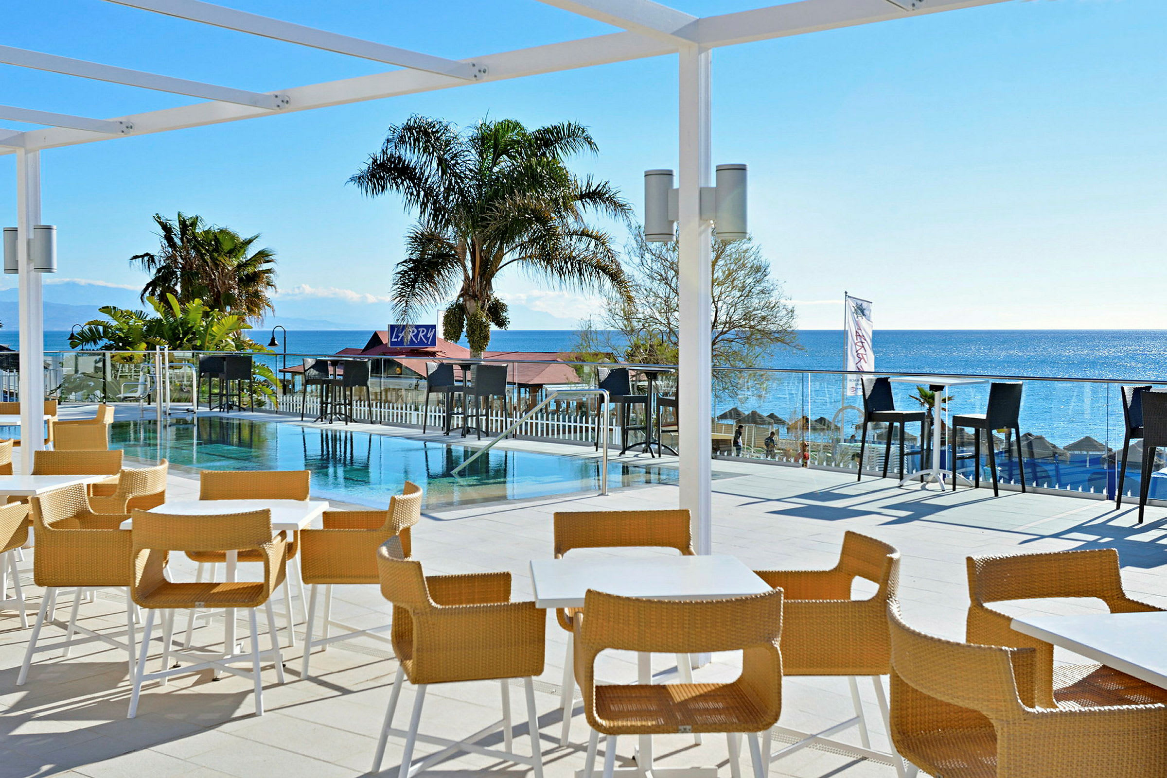Uteservering med bord och stolar vid en pool och havet i bakgrunden. (AI-genererad text)