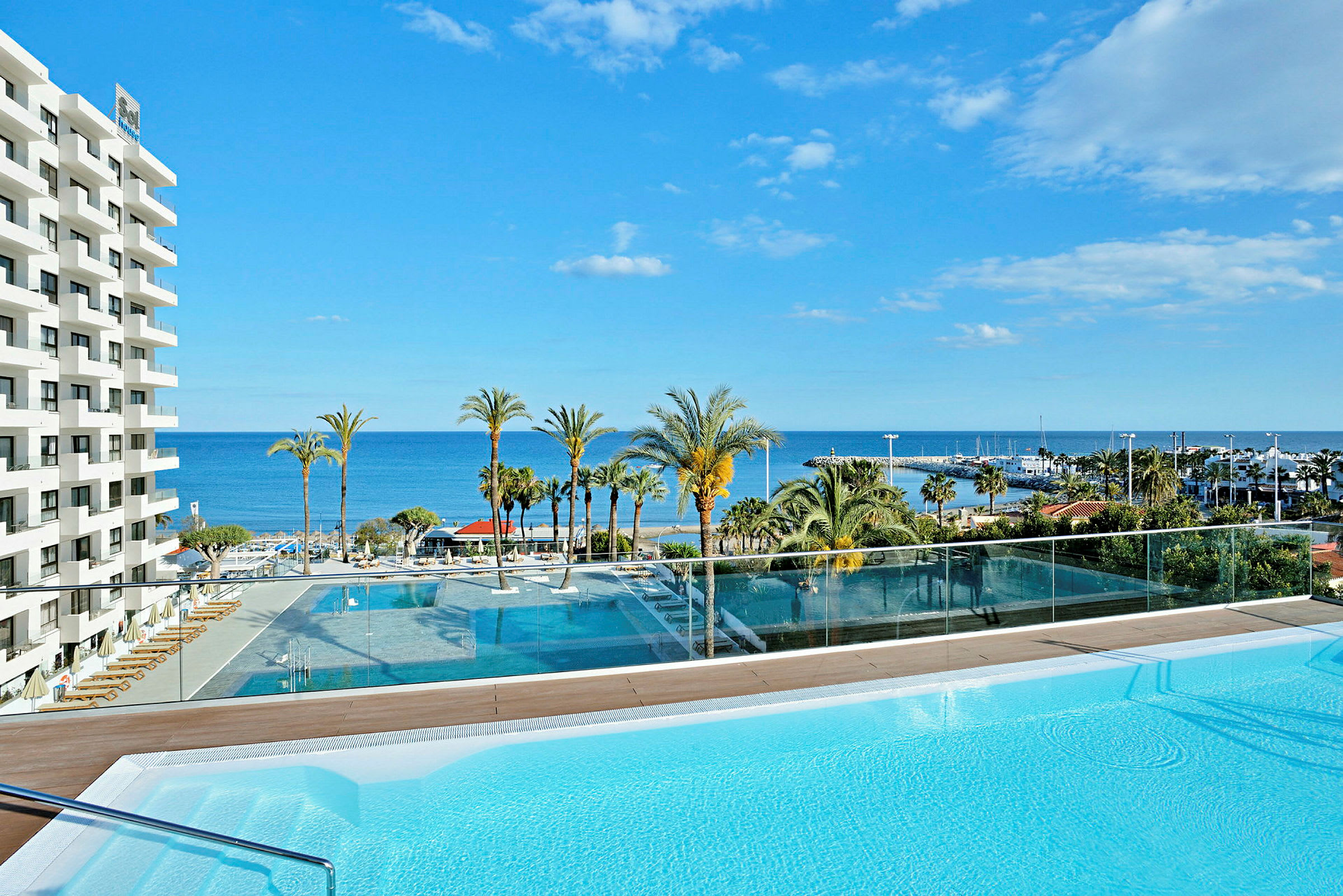 Utsikt över en swimmingpool med panoramavy mot havet och palmer samt en modern byggnad vid sidan. (AI-genererad text)