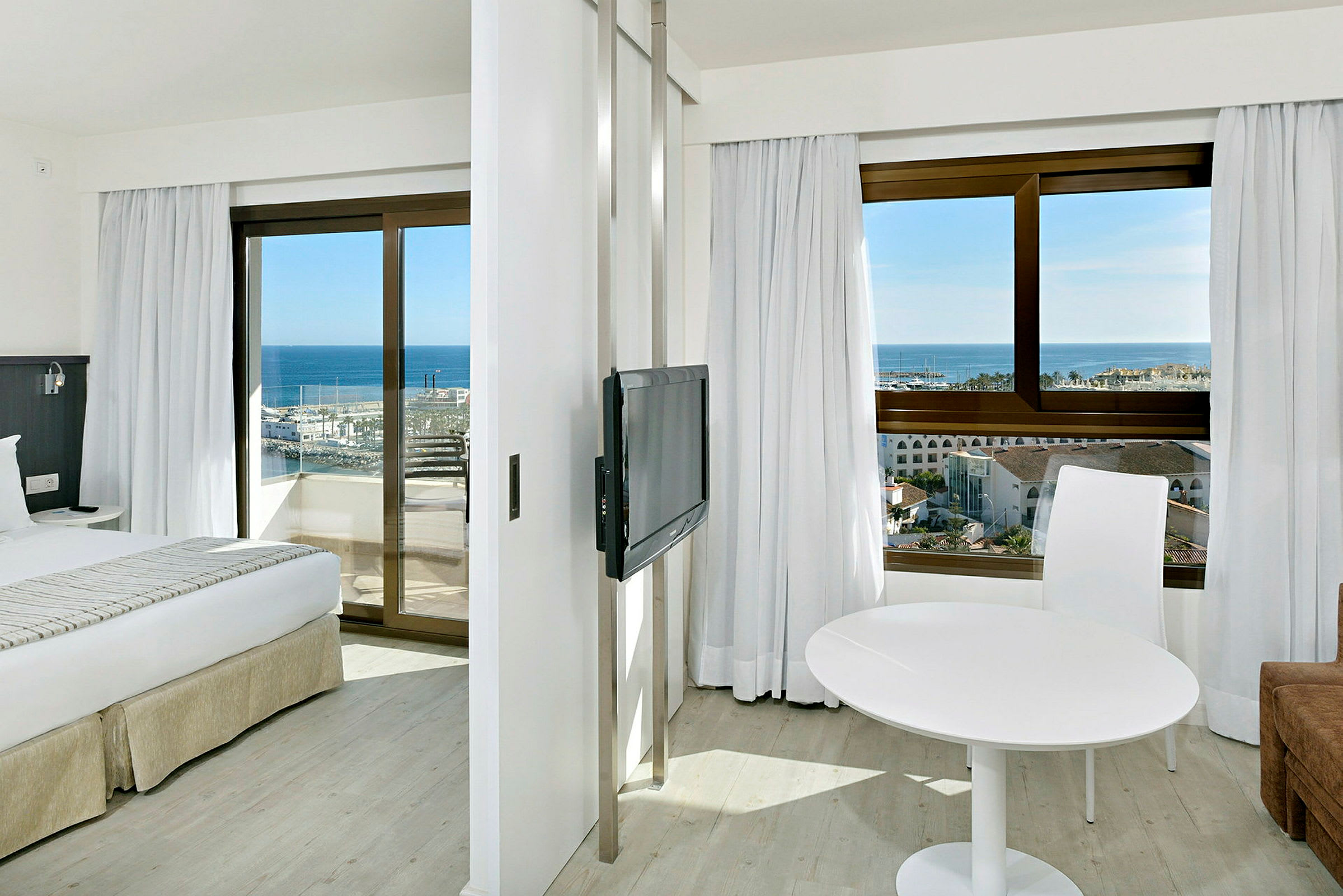 Hotellrum med två fönster som vetter mot havet, en säng, en TV och ett vitt bord med en stol. (AI-genererad text)