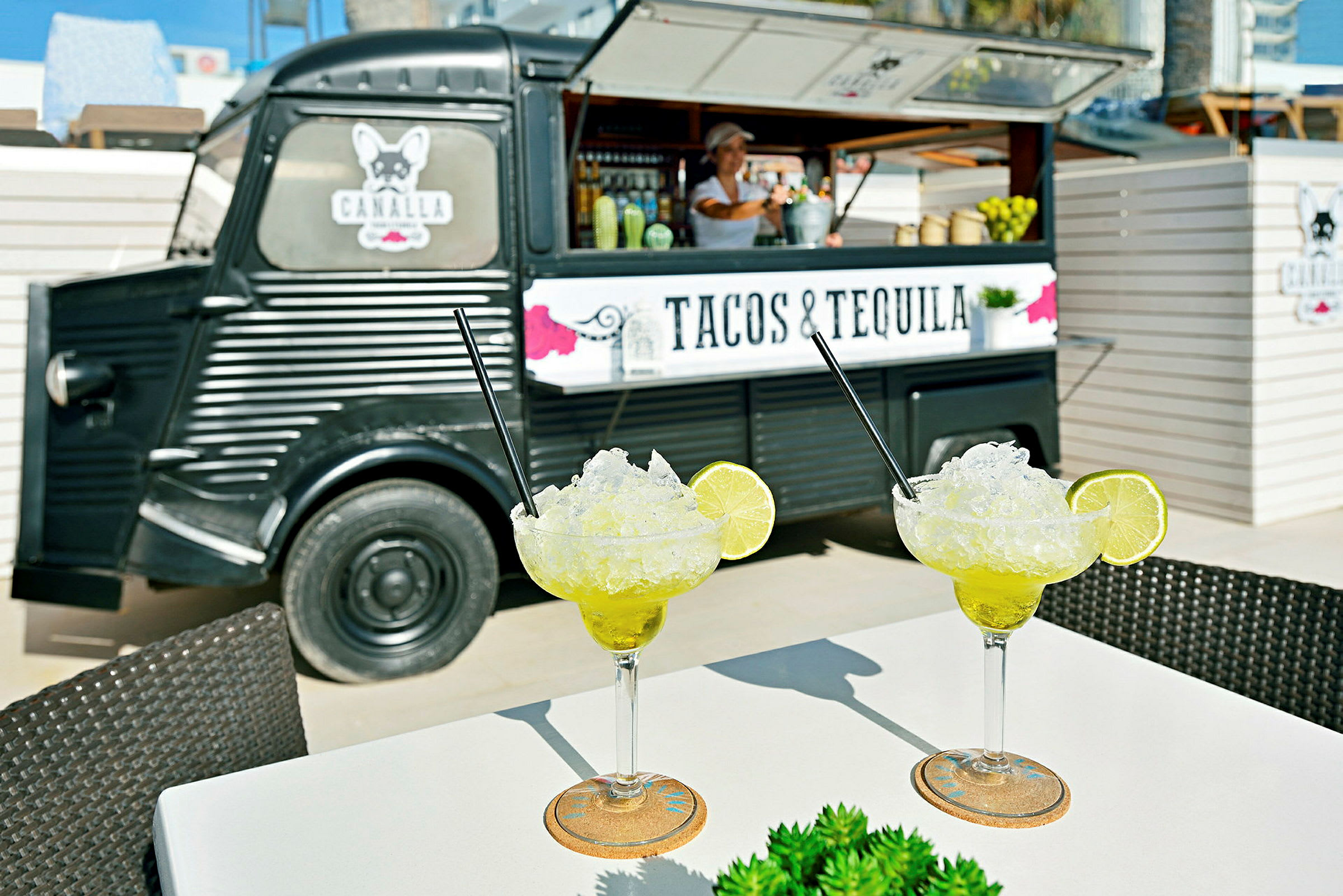 Två glas med margaritas står på ett bord framför en matbil med texten "Tacos & Tequila". (AI-genererad text)