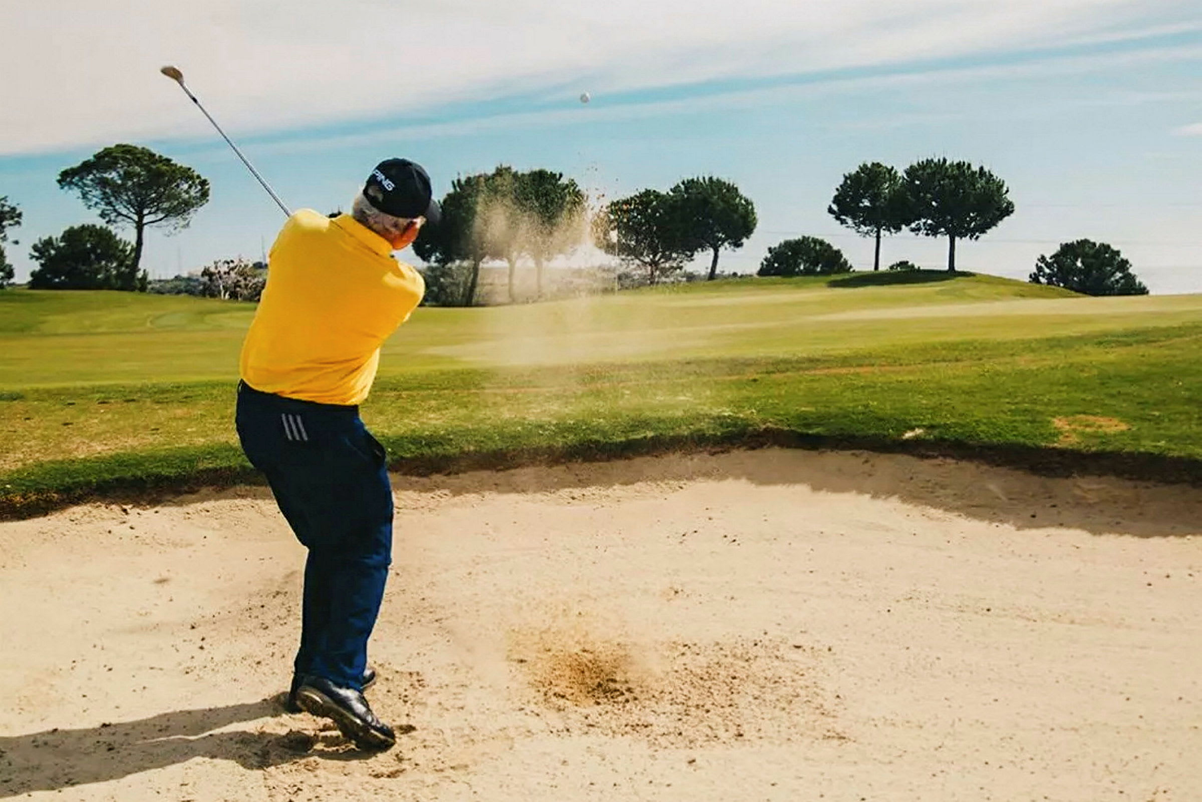 En person slår en golfboll från en sandbunker på en golfbana, omgiven av gröna träd. (AI-genererad text)