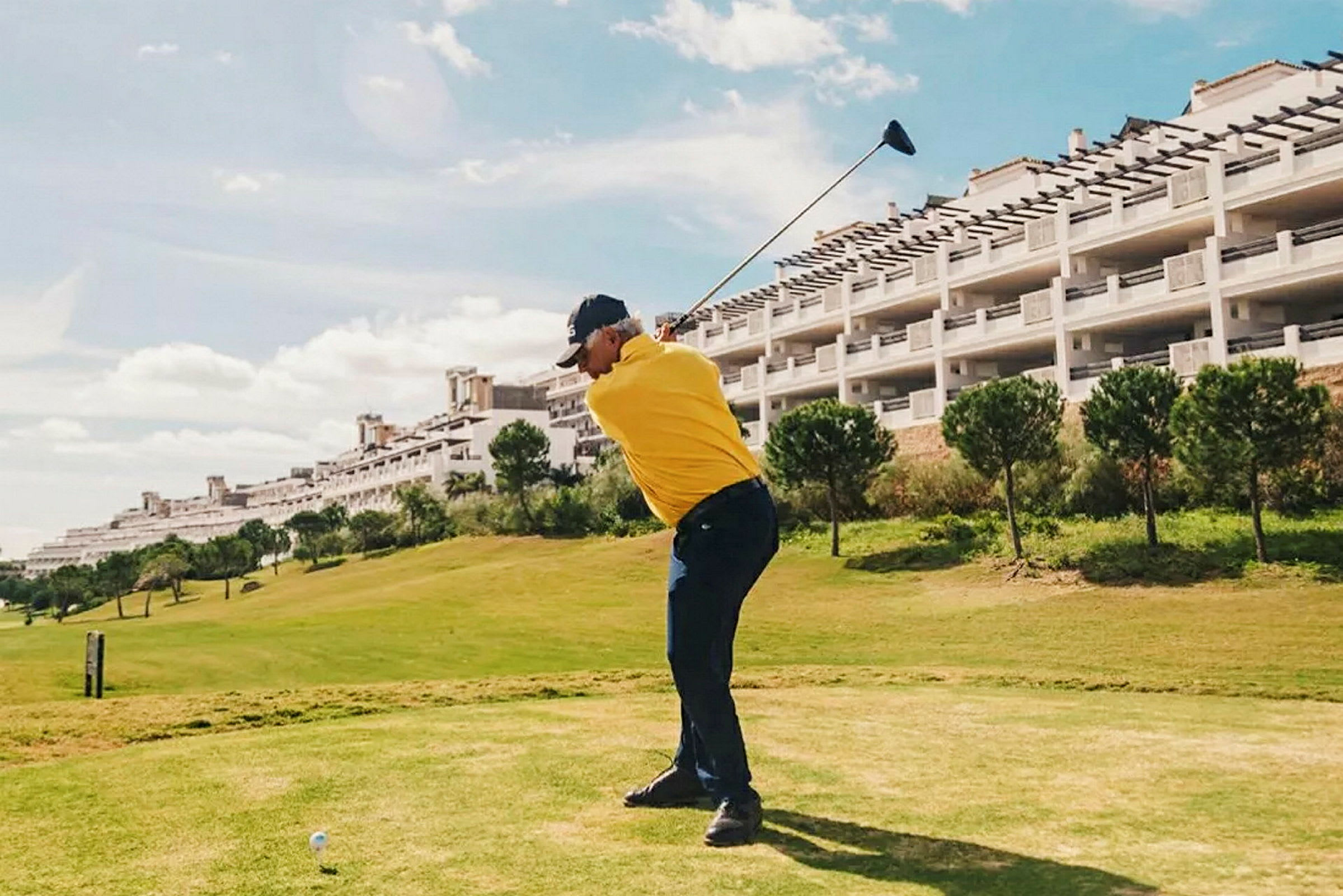 En person i gul tröja spelar golf på en grön bana med en byggnad och träd i bakgrunden. (AI-genererad text)