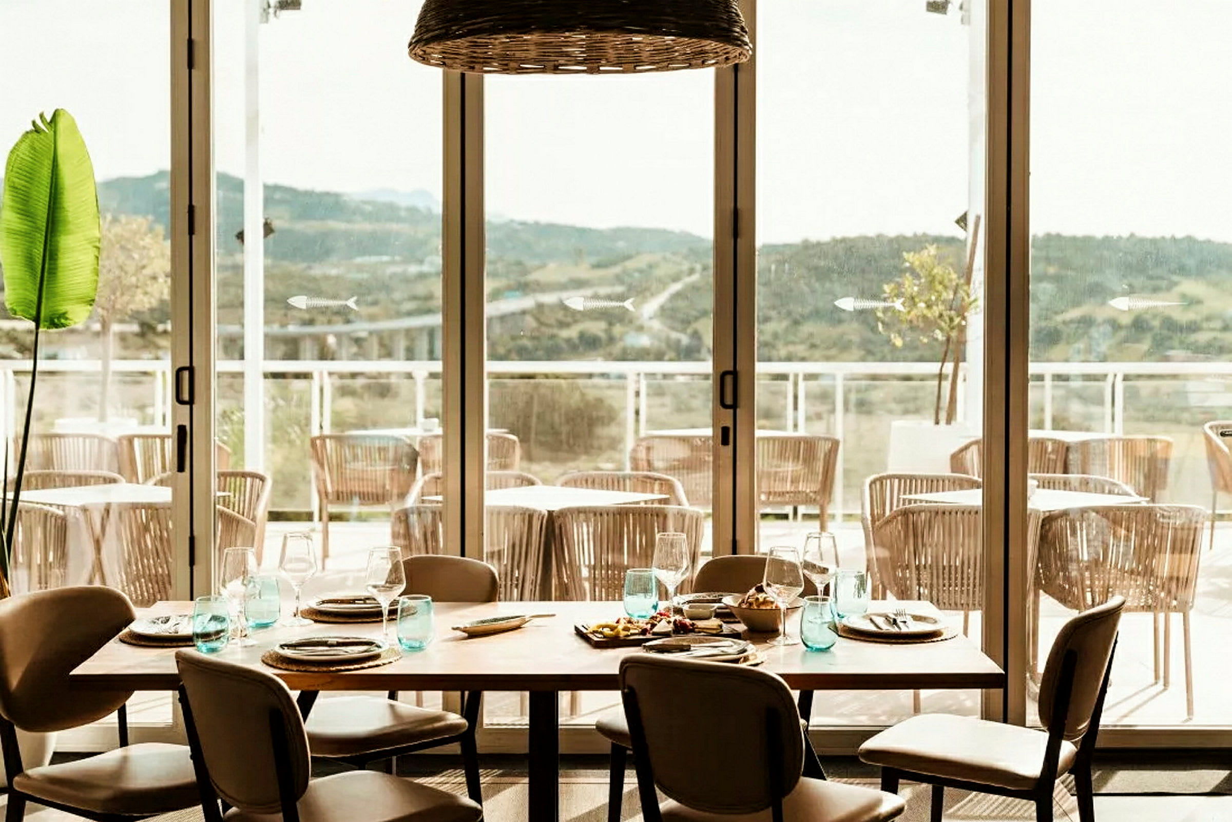 En restaurang med ett dukat bord och utsikt över en terrass med stolar och berg i fjärran. (AI-genererad text)