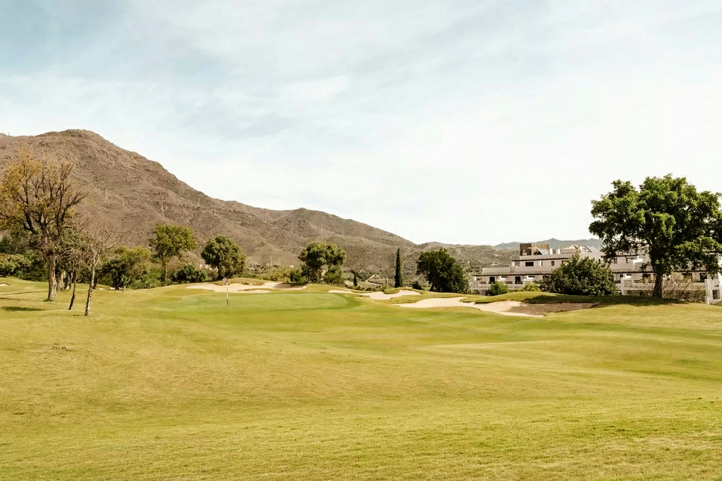 En golfbana omgiven av grön vegetation, med byggnader och berg i bakgrunden. (AI-genererad text)