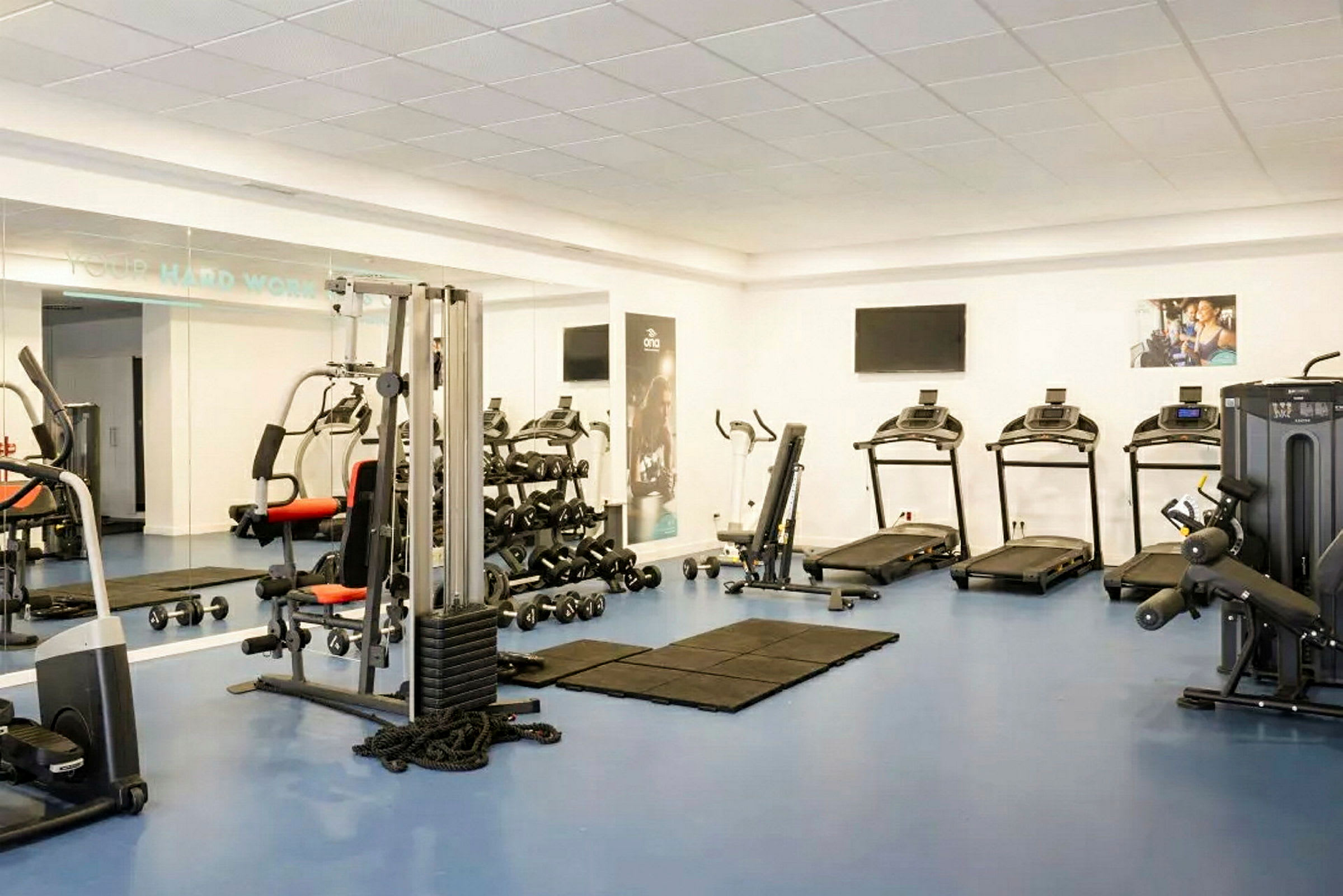 Ett gym med konditionsutrustning som löpband, träningsmaskiner och hantlar. Speglar täcker en vägg. (AI-genererad text)