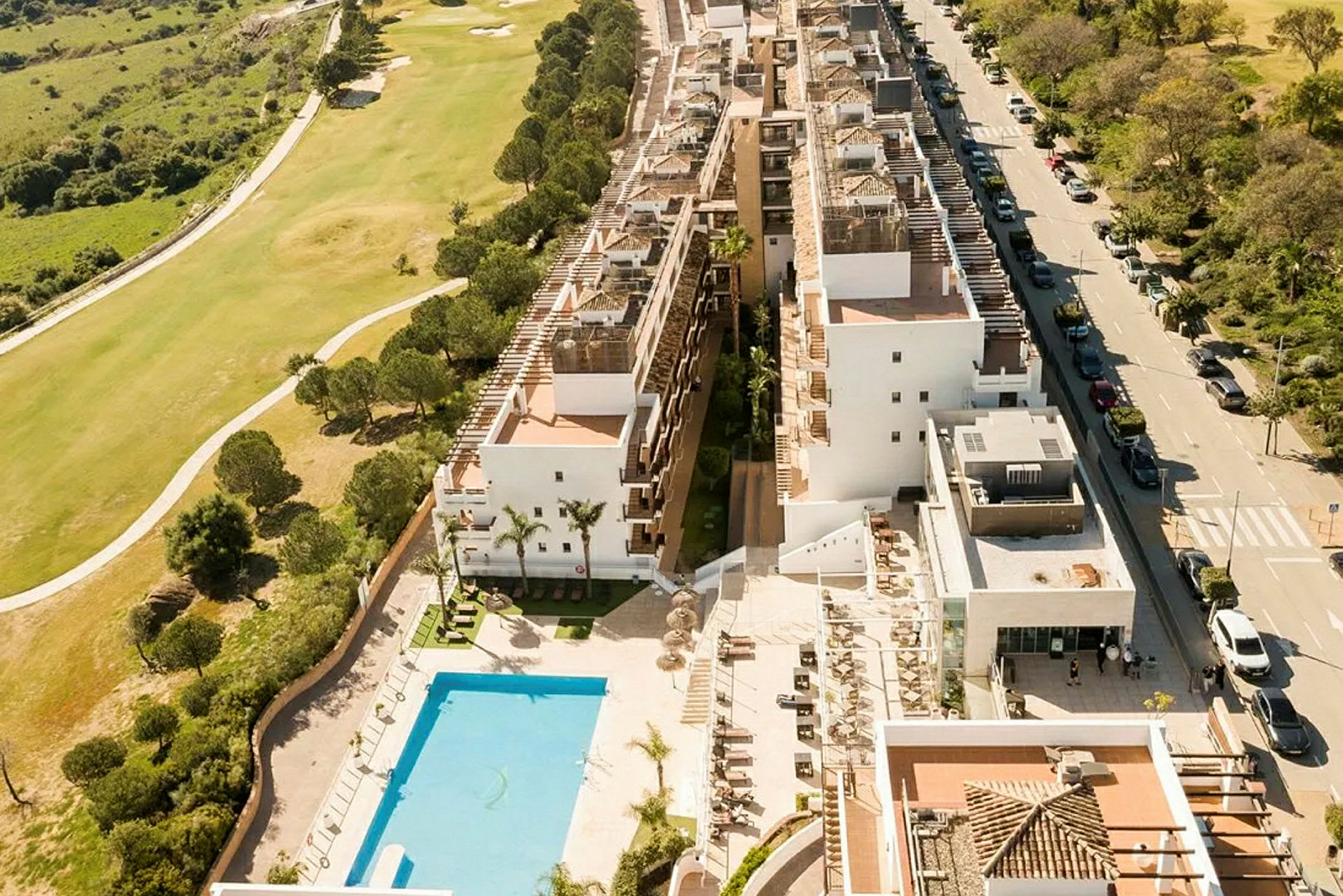 Flygfoto av en vit byggnadskomplex med poolområde och omgivande växtlighet. (AI-genererad text)