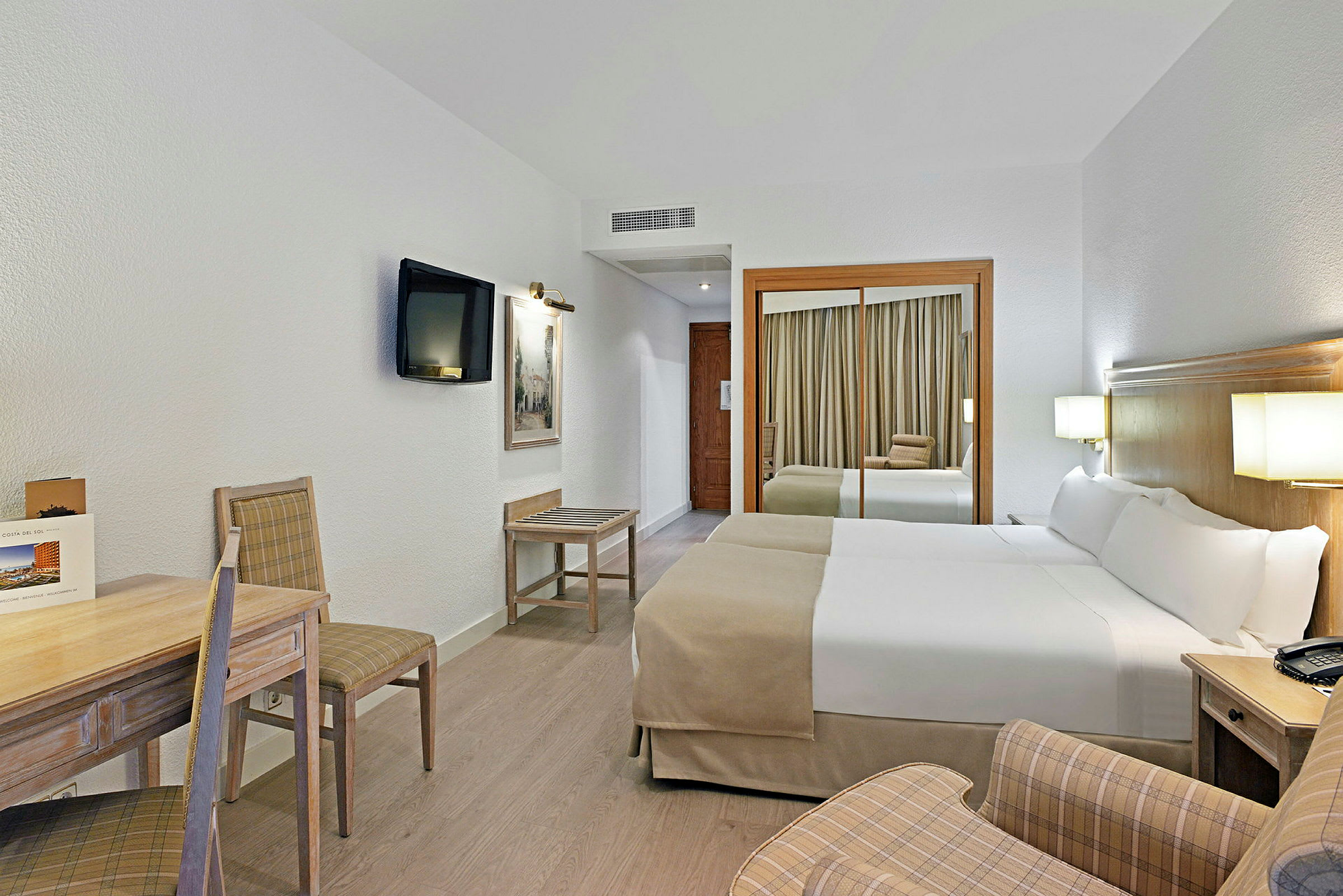 Ett hotellrum med två enkelsängar, en spegel, en tv och möbler. (AI-genererad text)