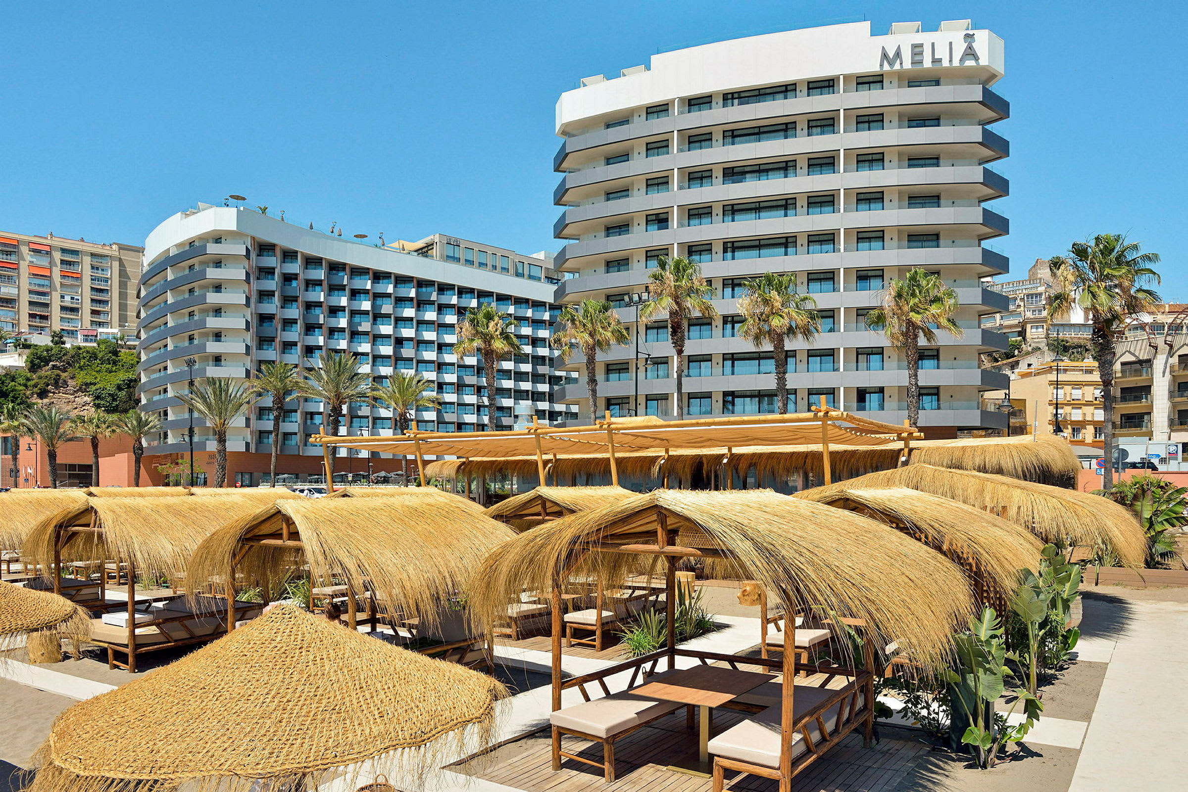 Ett hotell med flera våningar omgivet av palmer och strandpaviljonger med halmtak. (AI-genererad text)