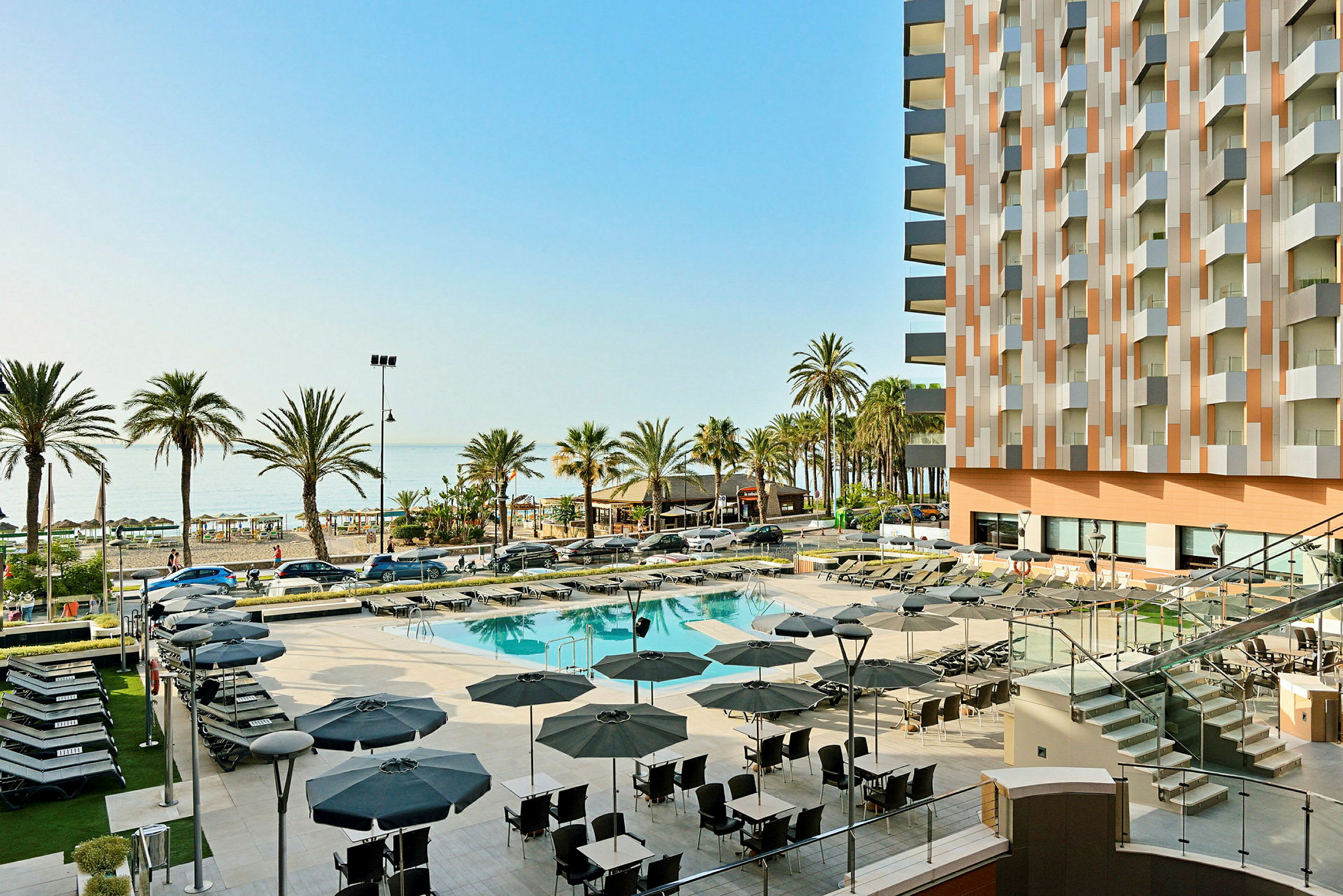 Bild av ett hotellområde med pool och solstolar, palmer i bakgrunden och havet synligt. (AI-genererad text)