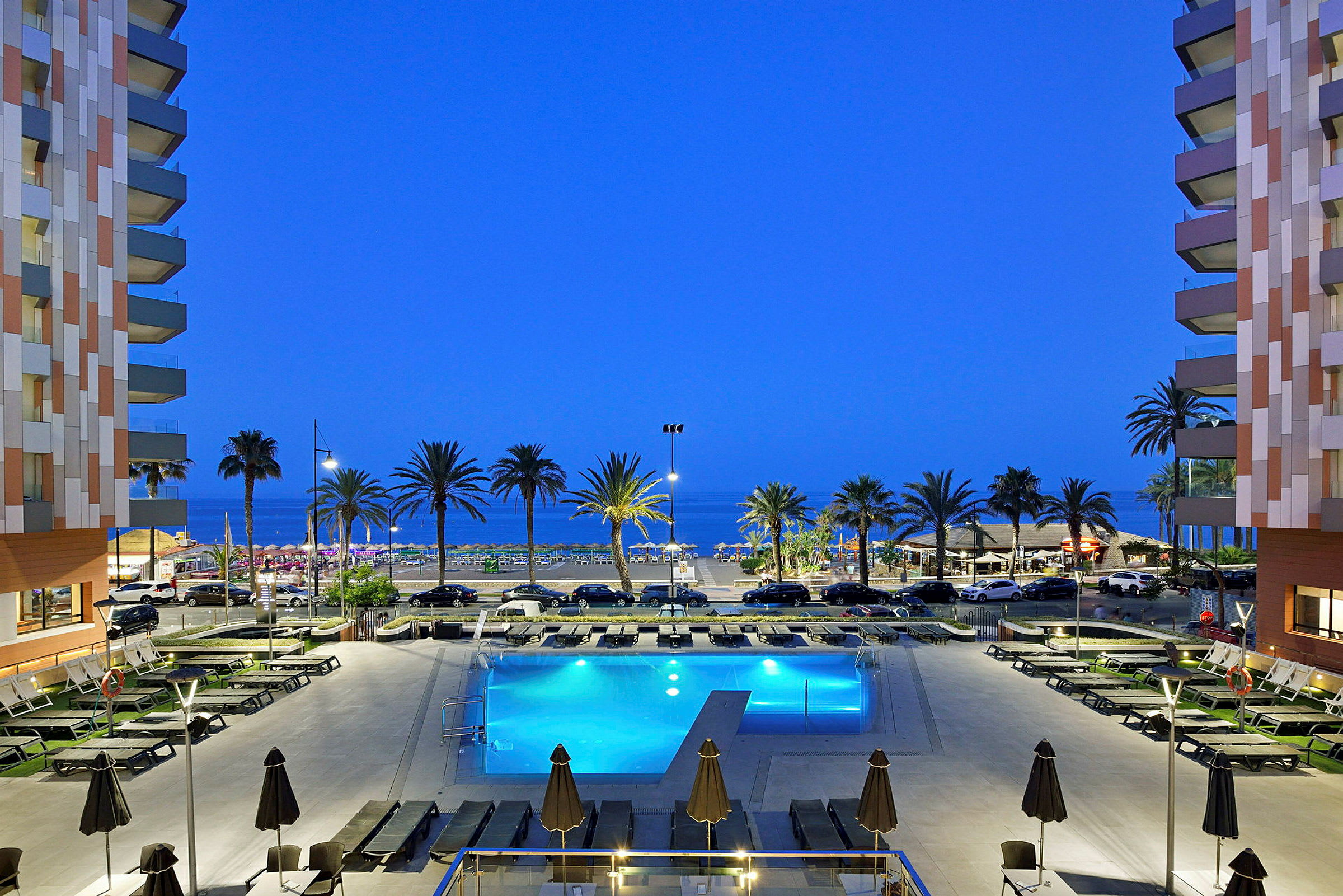 Bild av ett hotellområde med pool, solstolar och parasoller i förgrunden. I bakgrunden syns palmer, stranden och horisonten mot havet. (AI-genererad text)