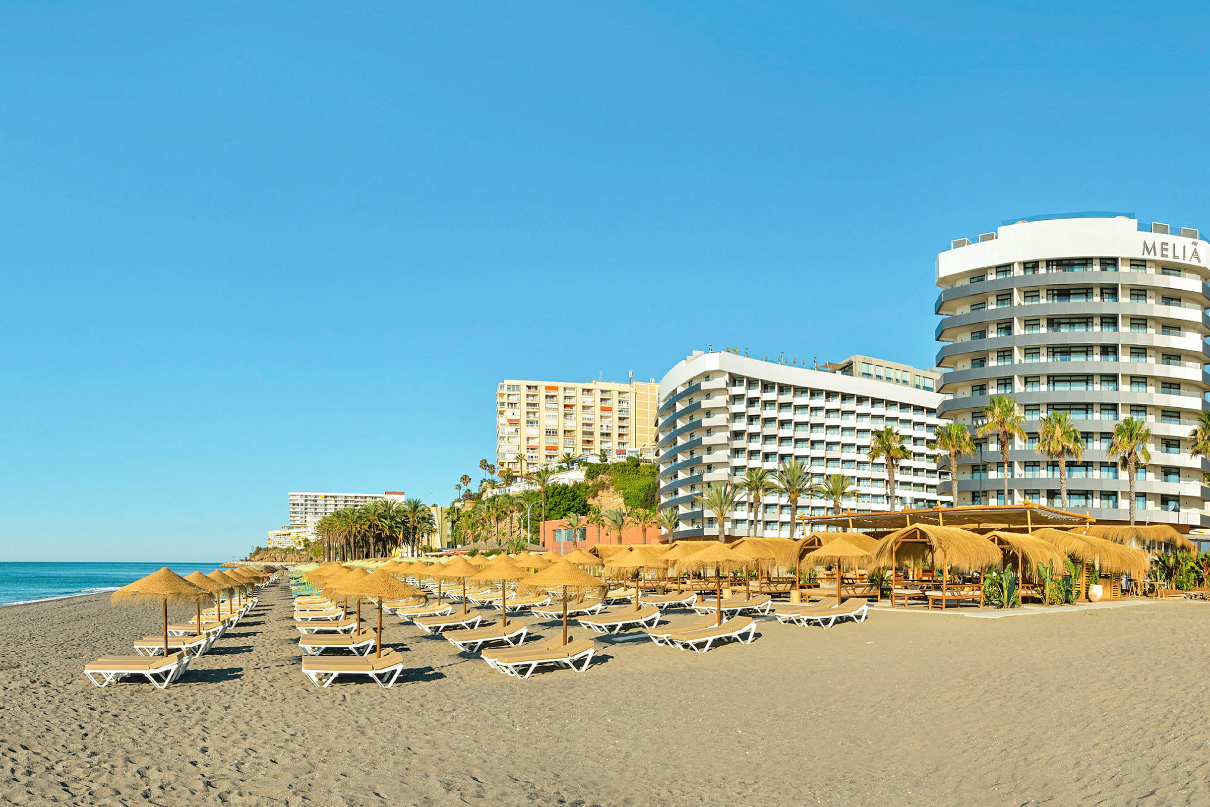 En strand med solstolar och parasoller, omgiven av flera hotellbyggnader. (AI-genererad text)