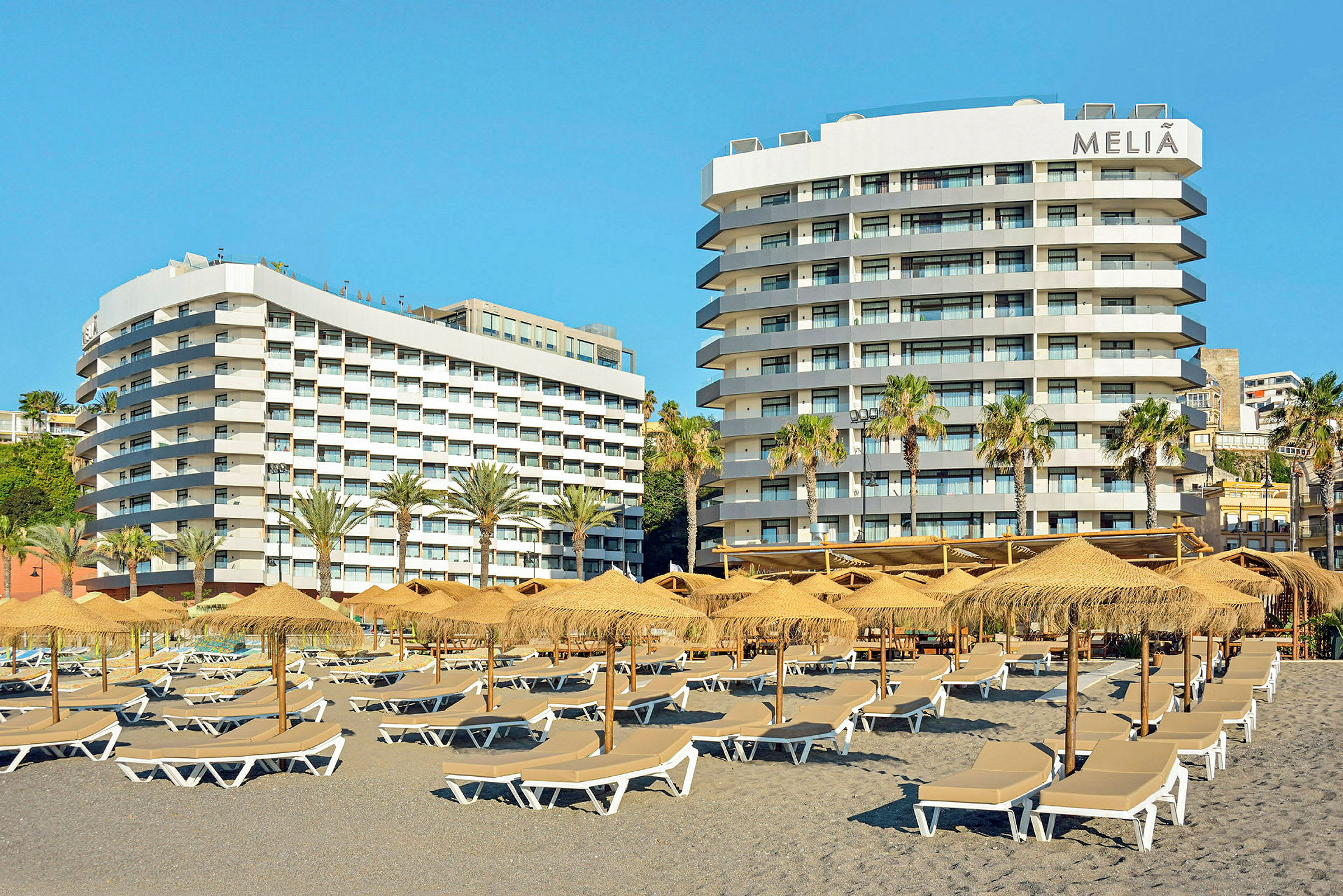 Bild av två hotellbyggnader med balkonger. Framför finns parasoll och solstolar på en strand. (AI-genererad text)