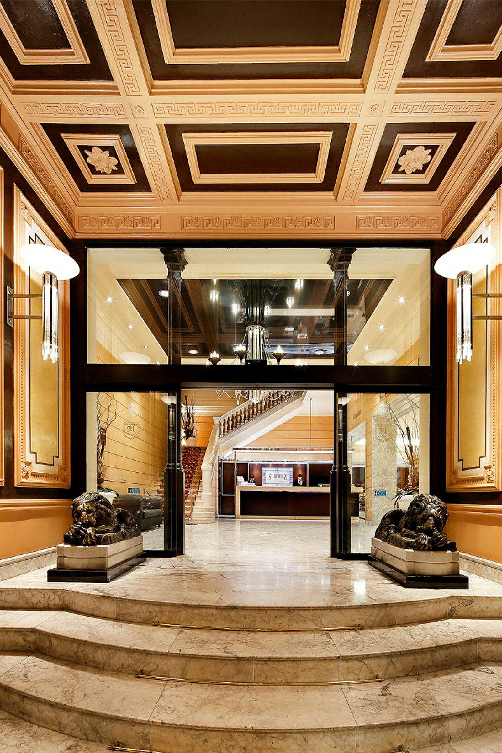 Bild av en elegant hotellfoajé med marmorgolv, utsmyckat tak och trappsteg som leder till receptionen. Två lejonstatyer flankerar entrén. (AI-genererad text)