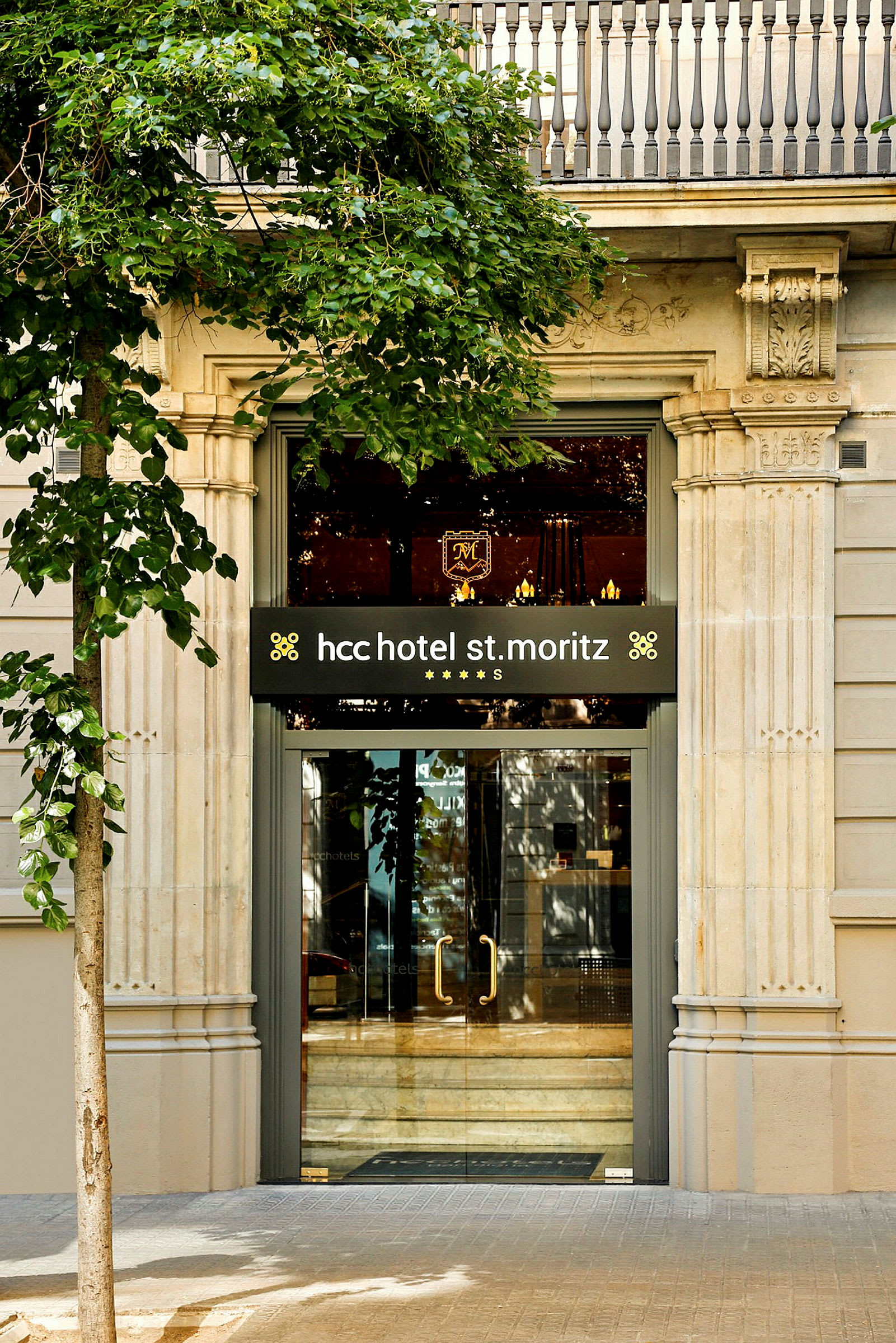 Entrén till hcc hotel St. Moritz, omgiven av grönska och en klassisk fasad. (AI-genererad text)