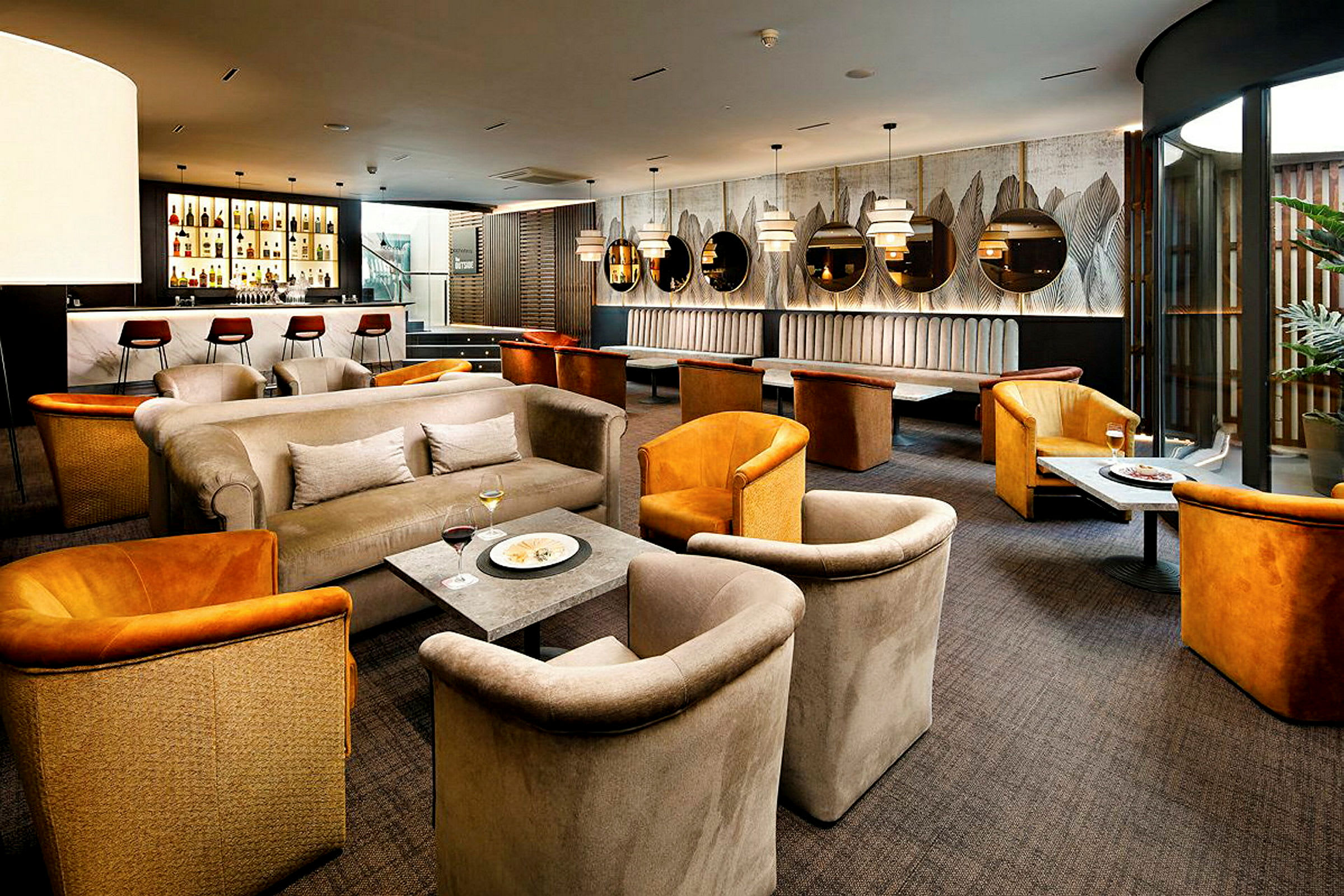 En stilfull lounge med soffor, fåtöljer och bord. Det finns en bardisk med barstolar i bakgrunden och väggdekorationer. (AI-genererad text)