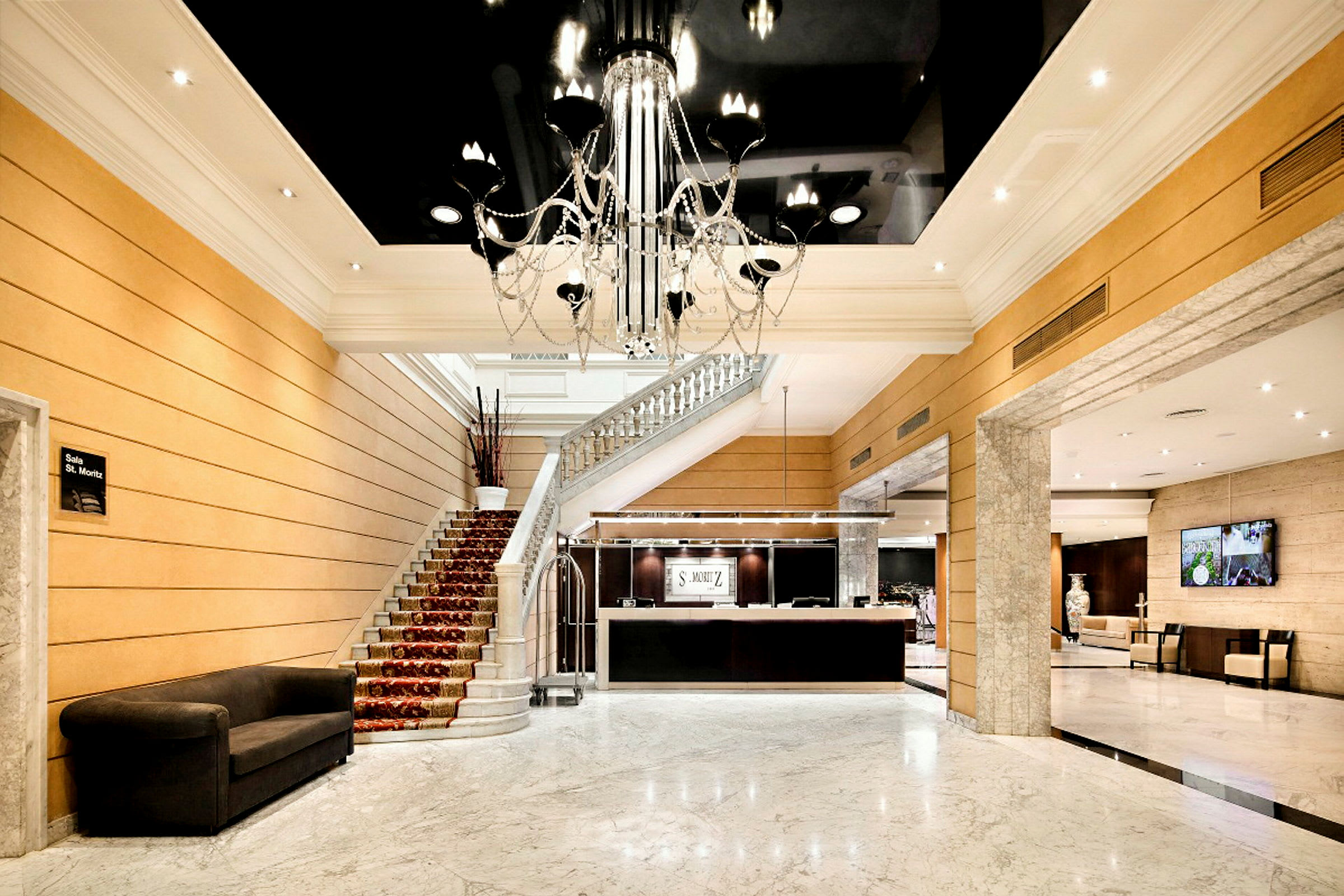 En hotellobby med marmorgolv och en elegant ljuskrona. En trappa leder upp från lobbyn och receptionen är synlig. (AI-genererad text)