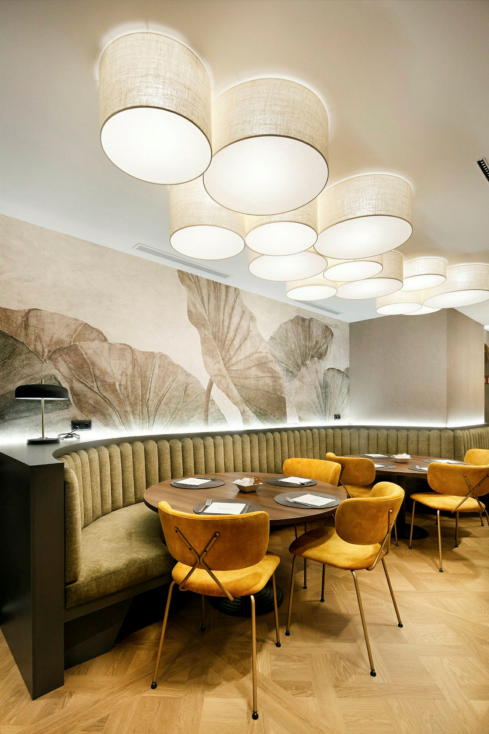 Interiör av en restaurang med runda träbord, gula stolar och en vägg med växtmotiv i bakgrunden. Taklampor med tygskärmar är monterade i taket. (AI-genererad text)