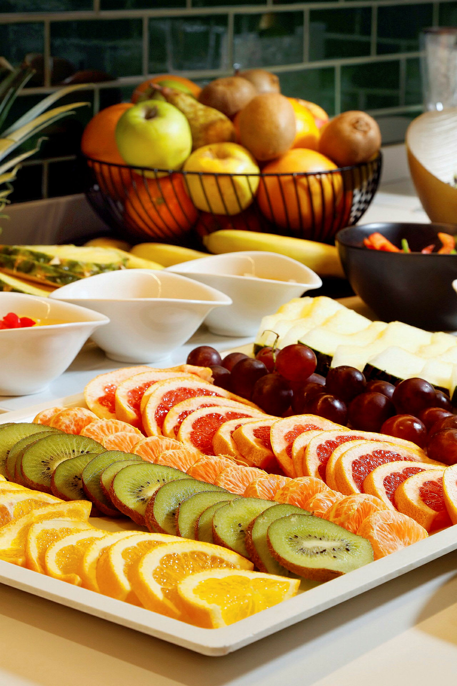 Ett uppläggningsfat med skivade frukter, inklusive kiwi, apelsin, och grapefrukt, samt hela vindruvor. Bakgrunden visar en skål med blandad frukt. (AI-genererad text)