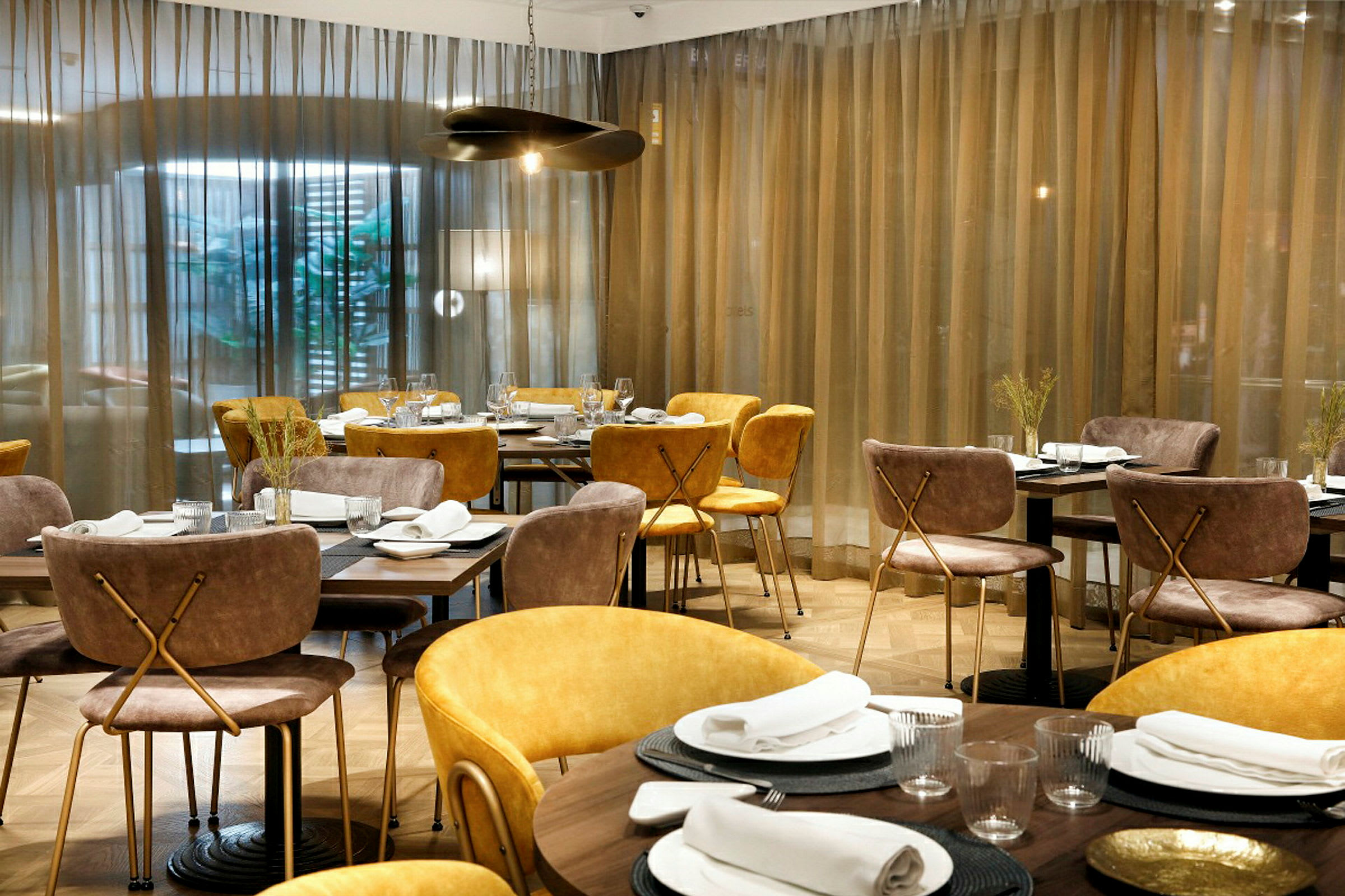 Ett elegant restaurangområde, med bord täckta av vita dukar och dukning. Stolarna är klädda i ljusbrunt och gult tyg, och gardiner pryder bakgrunden. (AI-genererad text)
