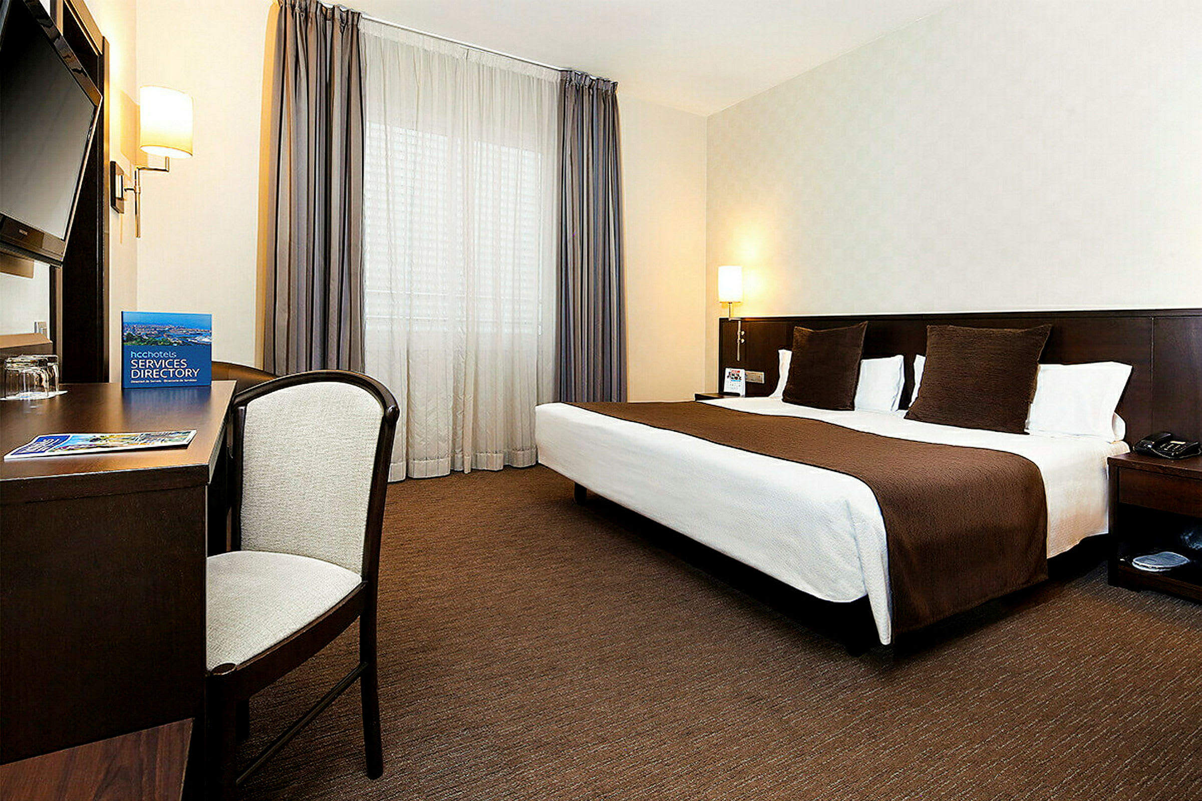 Ett hotellrum med en dubbelsäng, skrivbord, stol och stora fönster med gardiner. (AI-genererad text)