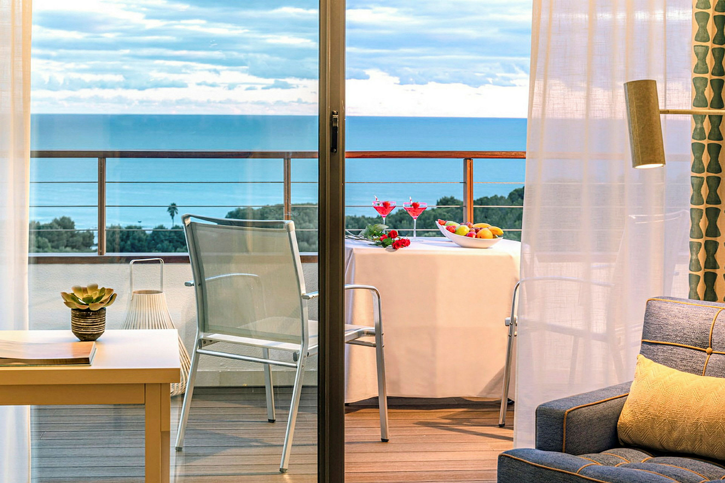 En balkong med utsikt över havet, ett bord med duk, frukt och drinkar, och stolar. Inomhus syns en soffa och dekoration. (AI-genererad text)