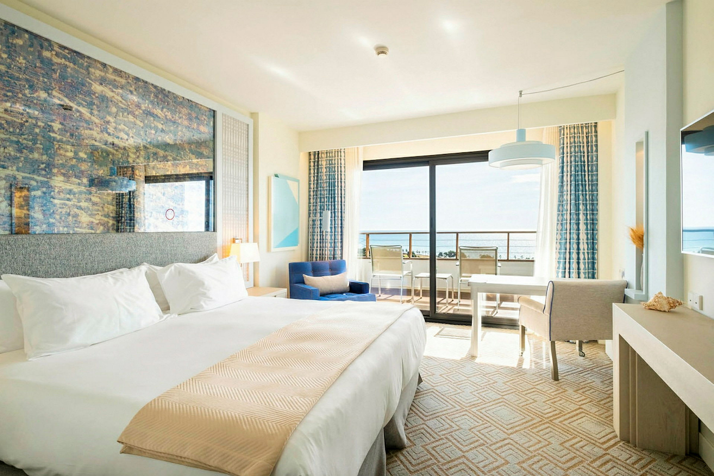 Ett hotellrum med en stor säng, sittgrupp och skrivbord. Det finns en balkong med utsikt över havet. (AI-genererad text)