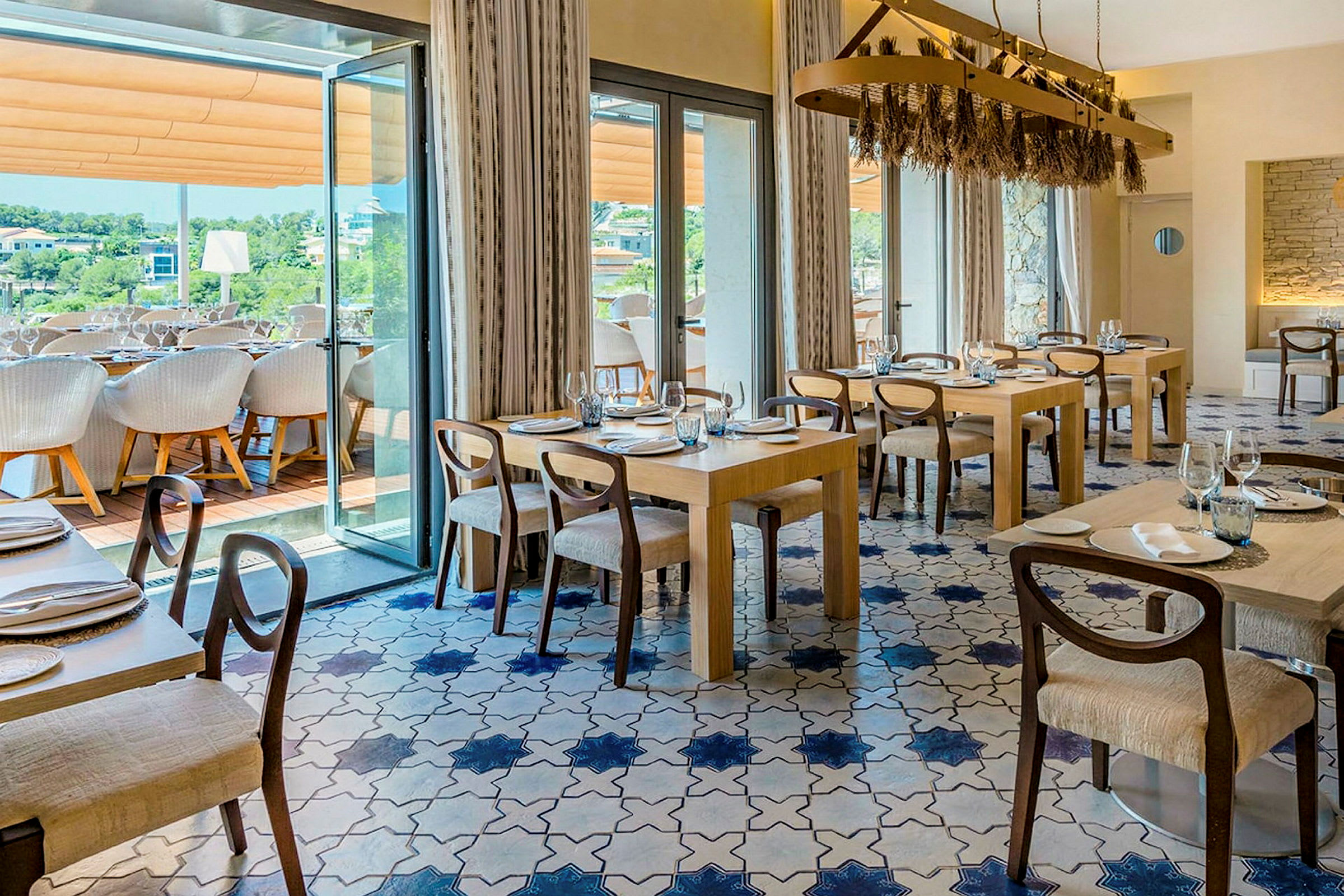 En elegant restaurang med flera bord och stolar, blått och vitt kakelgolv och stora fönster som leder ut till en terrass. (AI-genererad text)