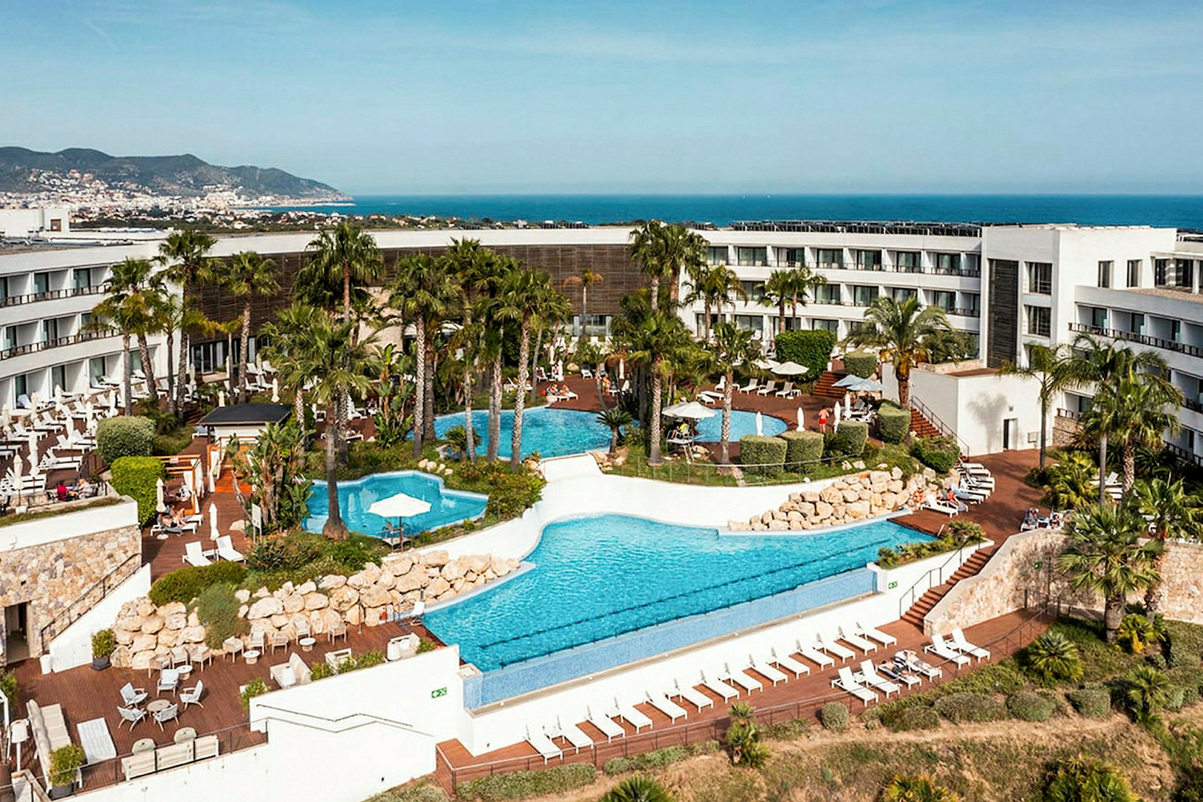 Ett hotell med pool område och omgivande trädgård, med havet och en stad i bakgrunden. (AI-genererad text)