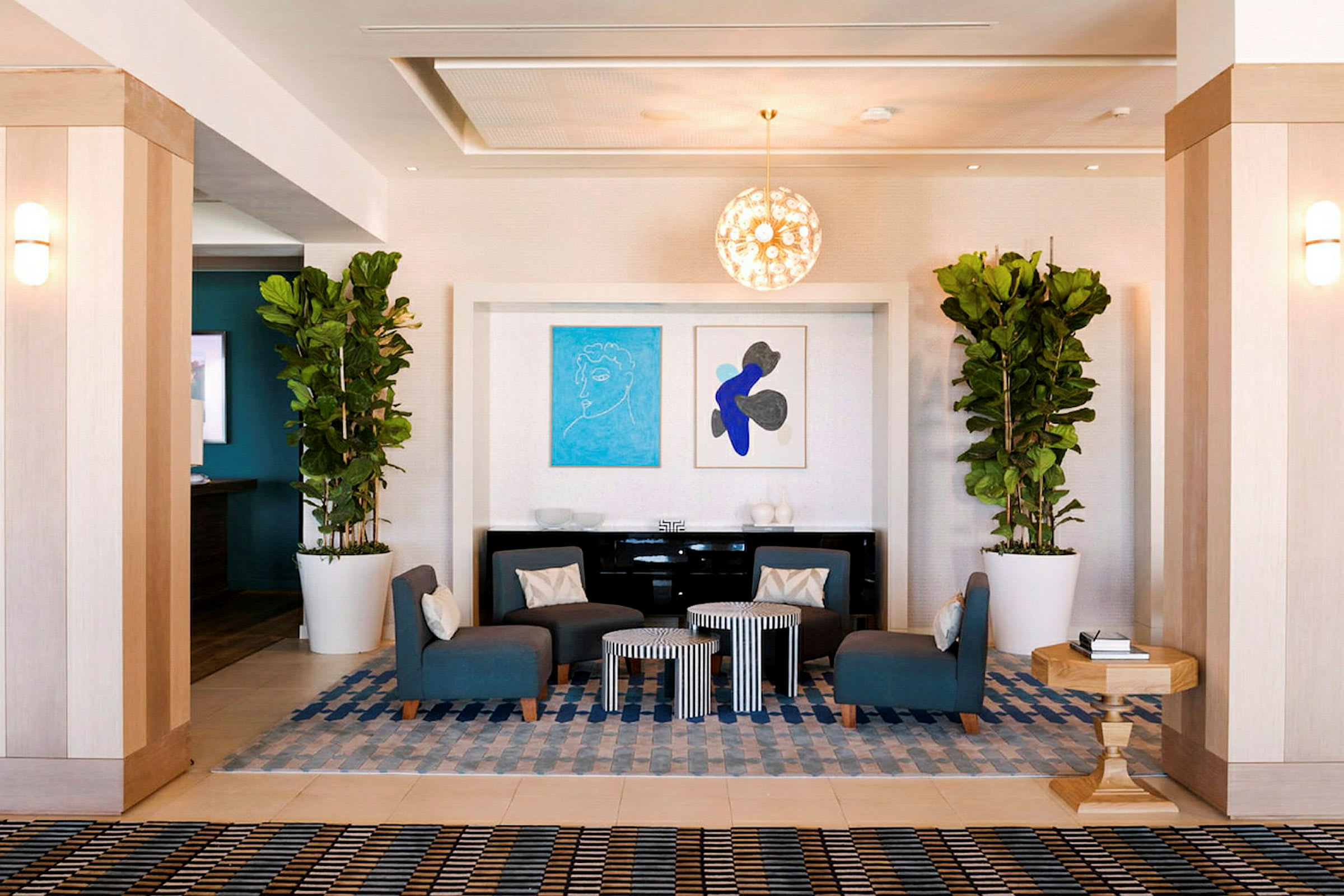 En hotellobby med sittplatser i form av blå fåtöljer runt små bord. Växter i stora vita krukor och konstverk på väggen dekorerar rummet. (AI-genererad text)