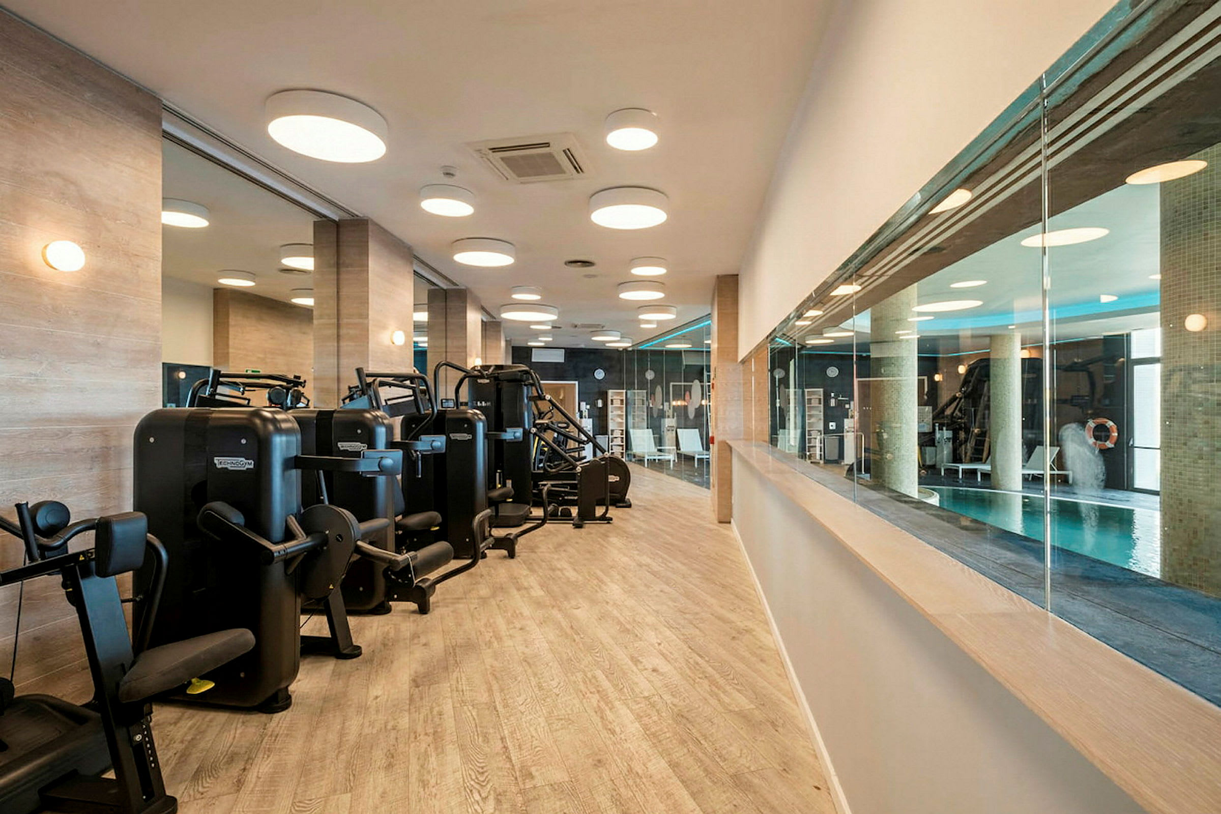 Ett gym med träningsmaskiner och speglar på väggarna, samt en glasvägg som vetter mot en inomhuspool. (AI-genererad text)