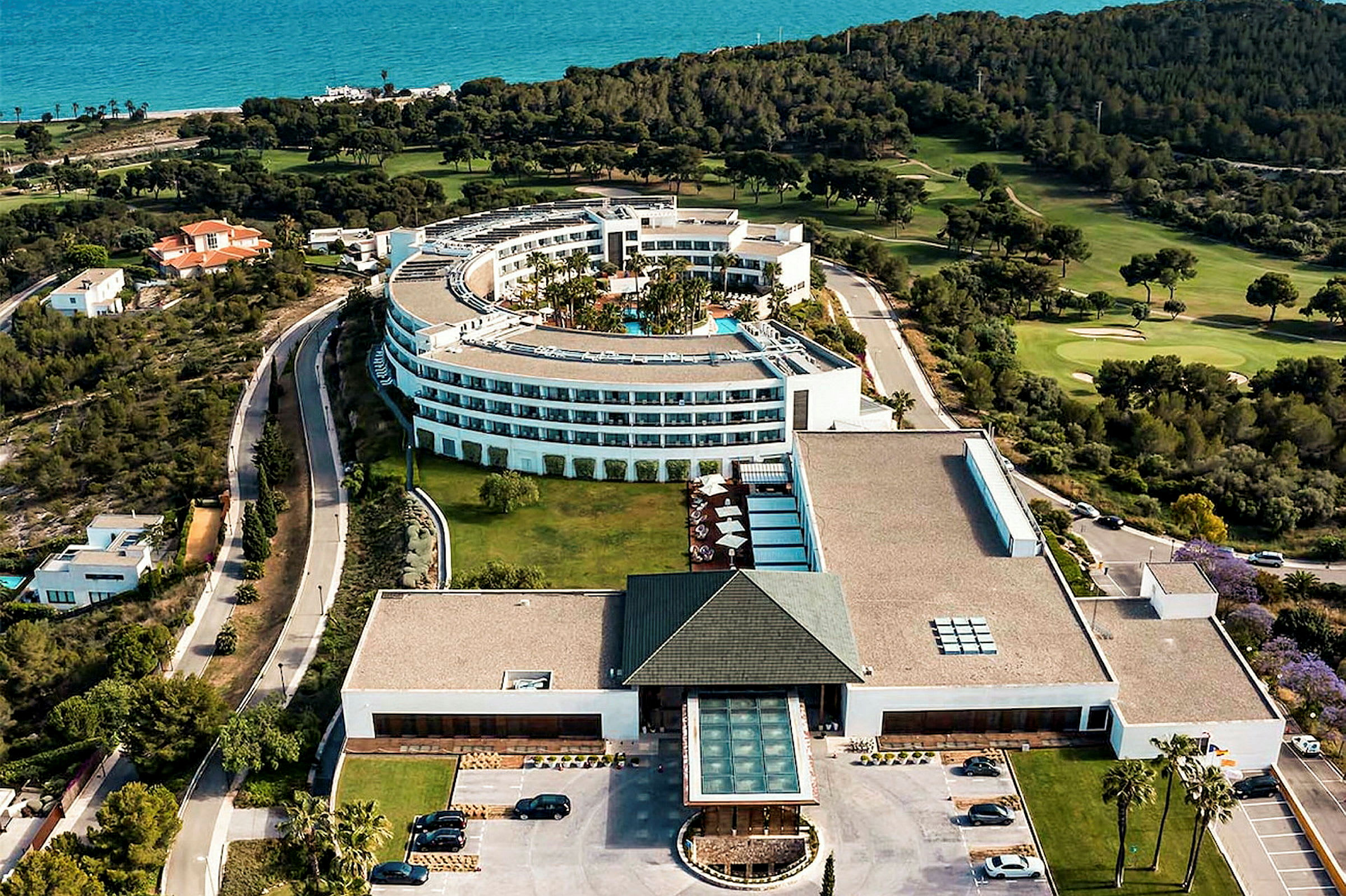 Flygfoto av en stor vit hotellbyggnad med en modern design, omgiven av grönska och nära havet. (AI-genererad text)